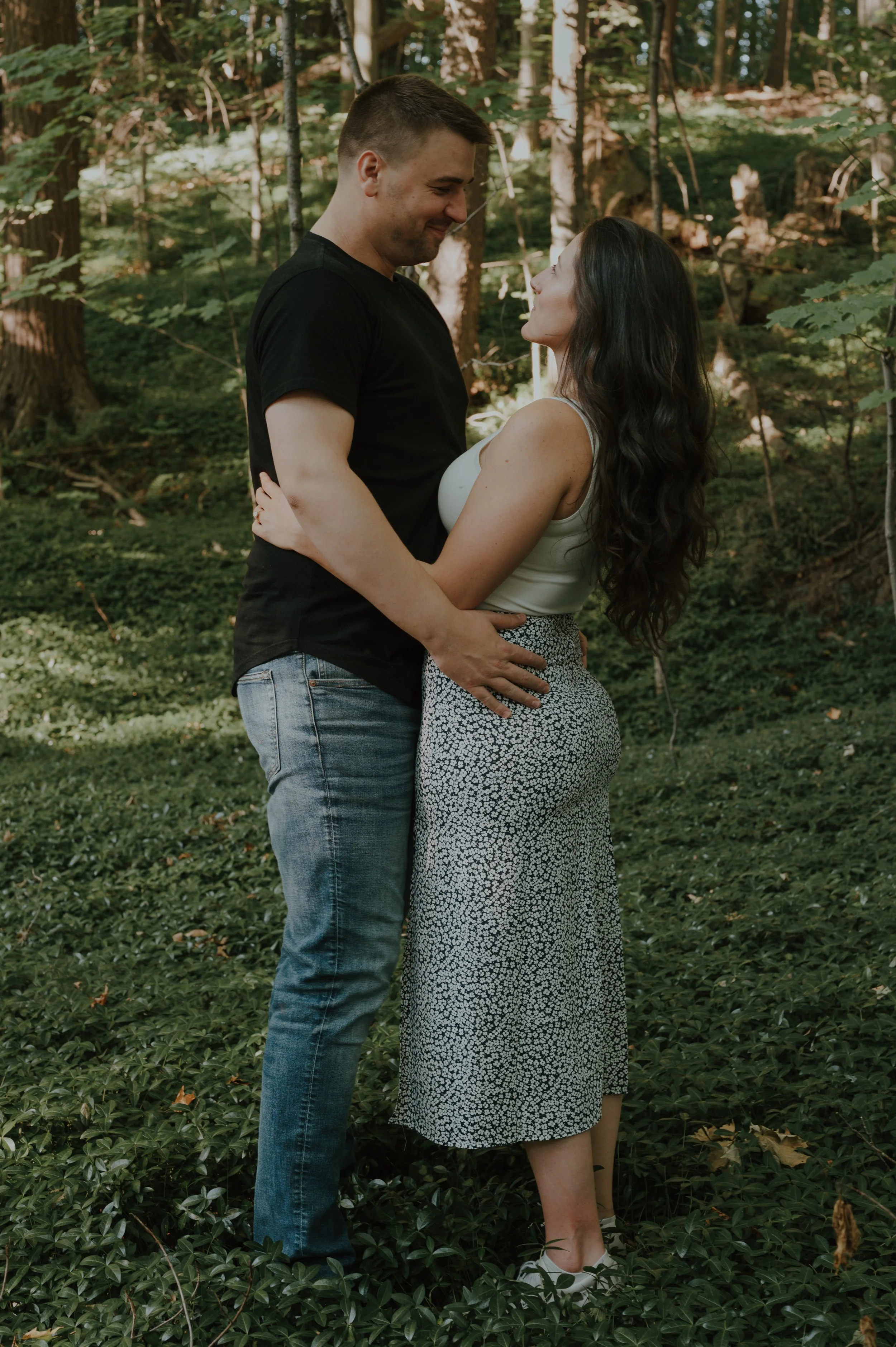 A couple stands close, embracing and looking into each other's eyes in a forested area.