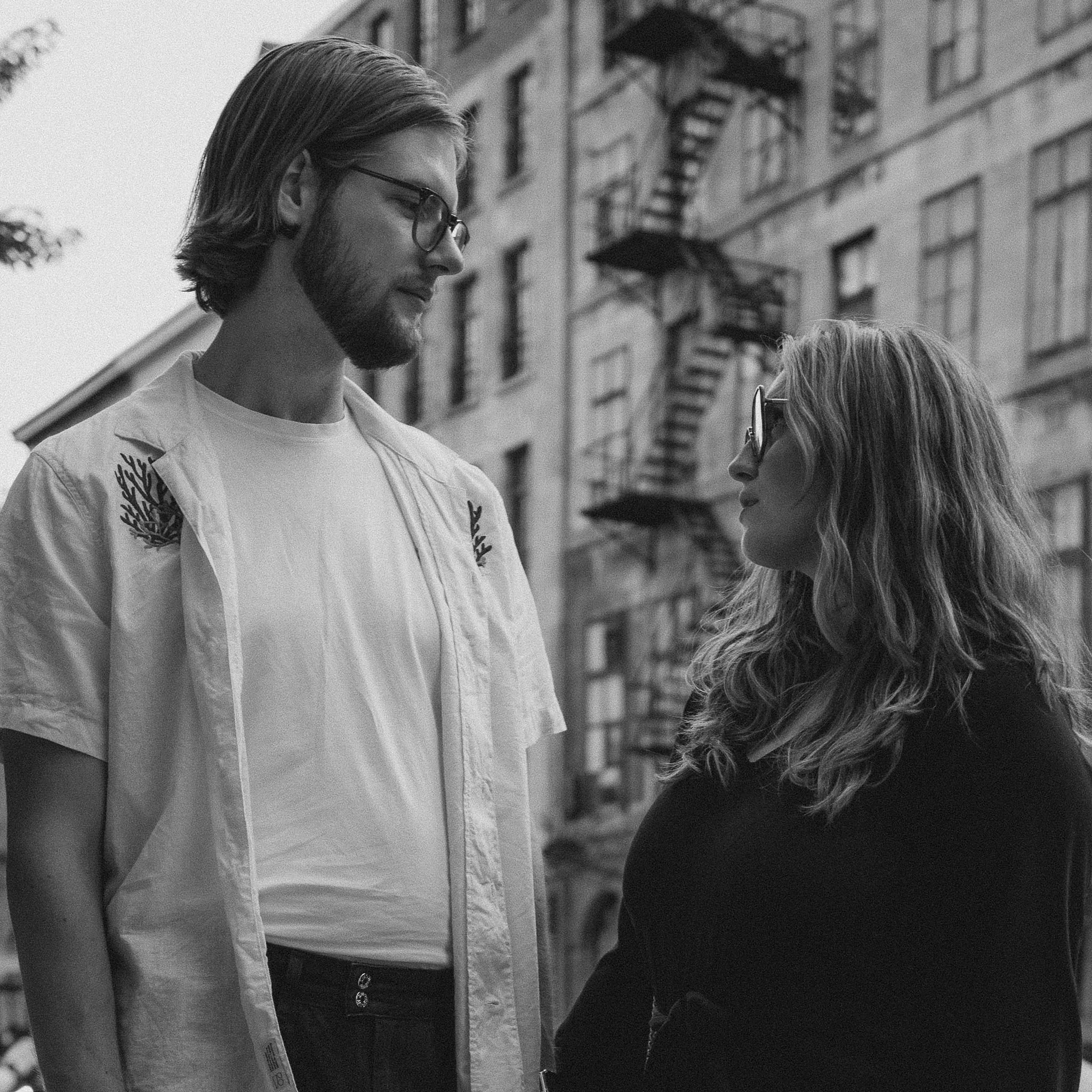 A black and white photo of a man and woman standing face to face outdoors in an urban setting, with a building that has fire escape stairs in the background. The man has long hair, a beard, and glasses, and is wearing a casual shirt with embroidered 