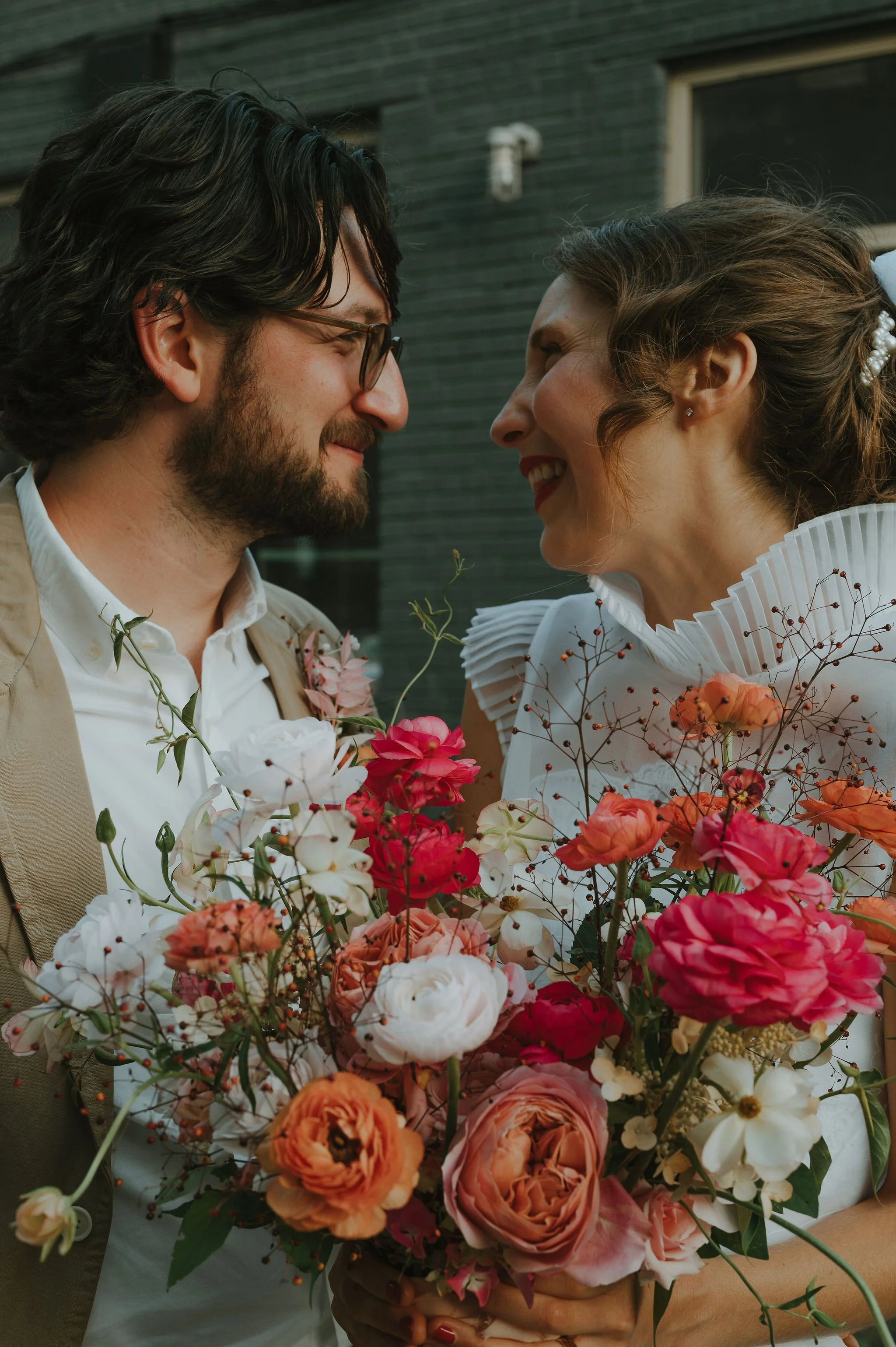 A man and woman smiling close together, holding a large bouquet of colorful flowers, outside in front of a dark building.