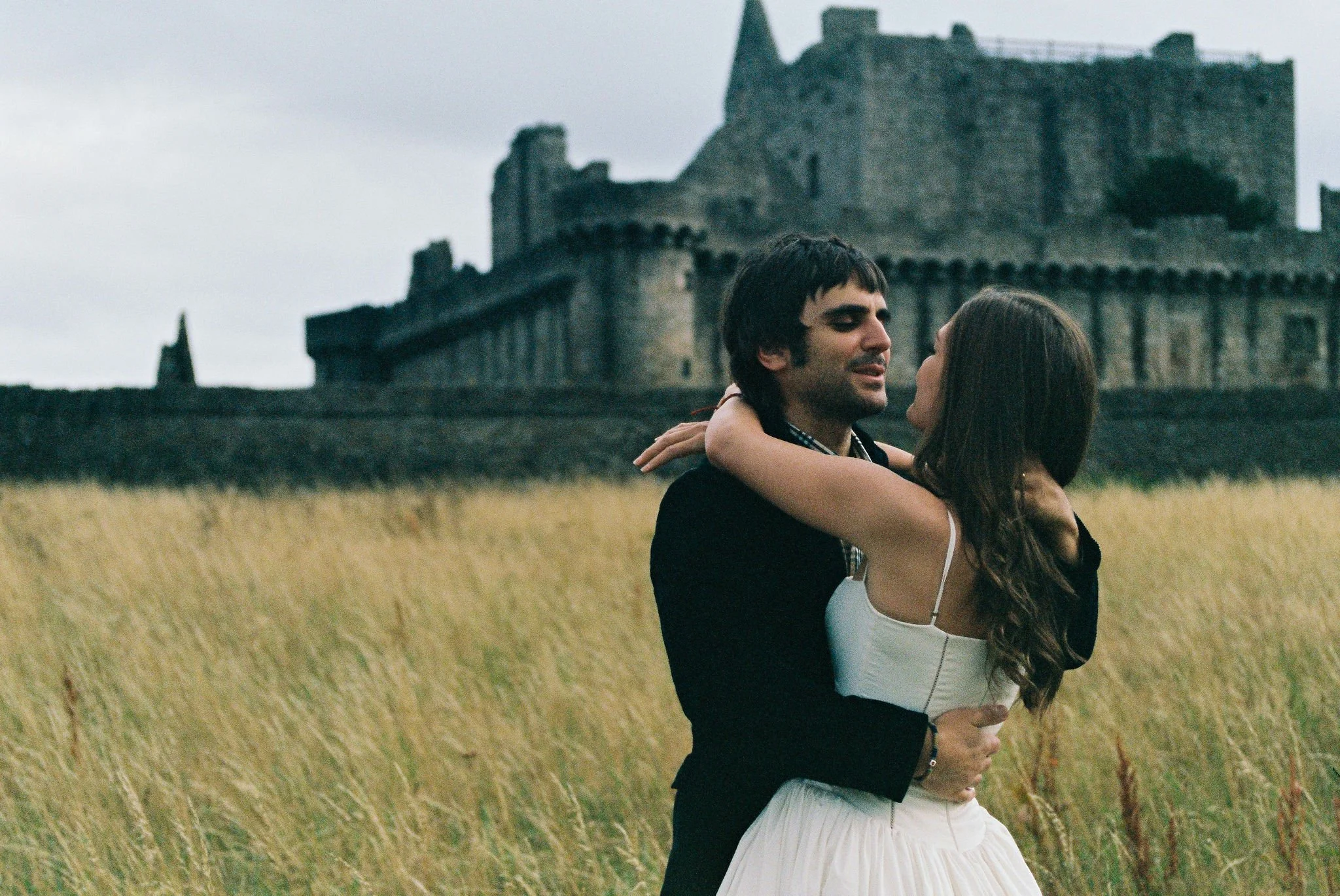 A couple dancing in a field near a castle on a cloudy day.