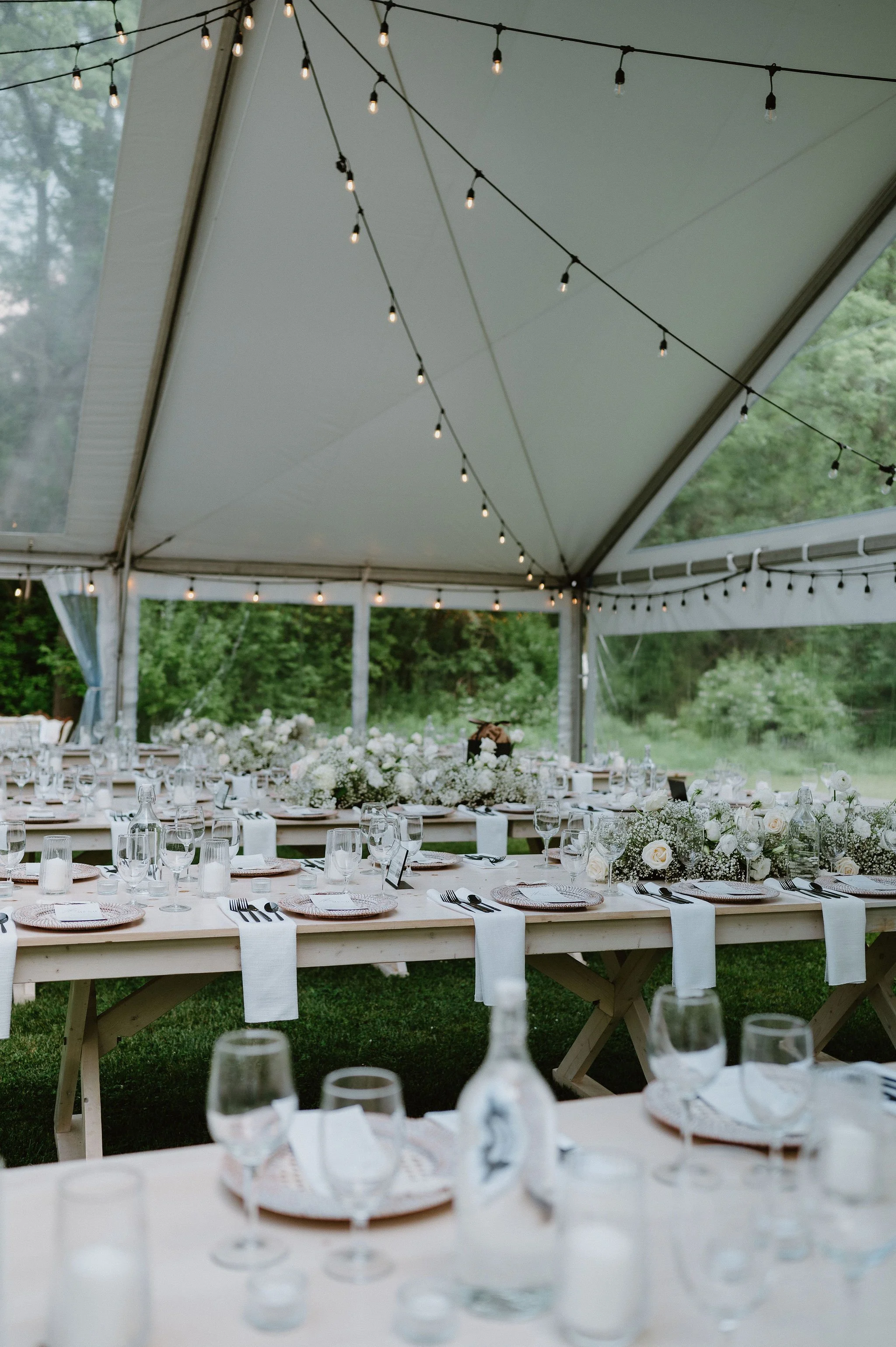 Wedding reception setup with long tables, white flowers, and tableware under a white tent decorated with string lights.