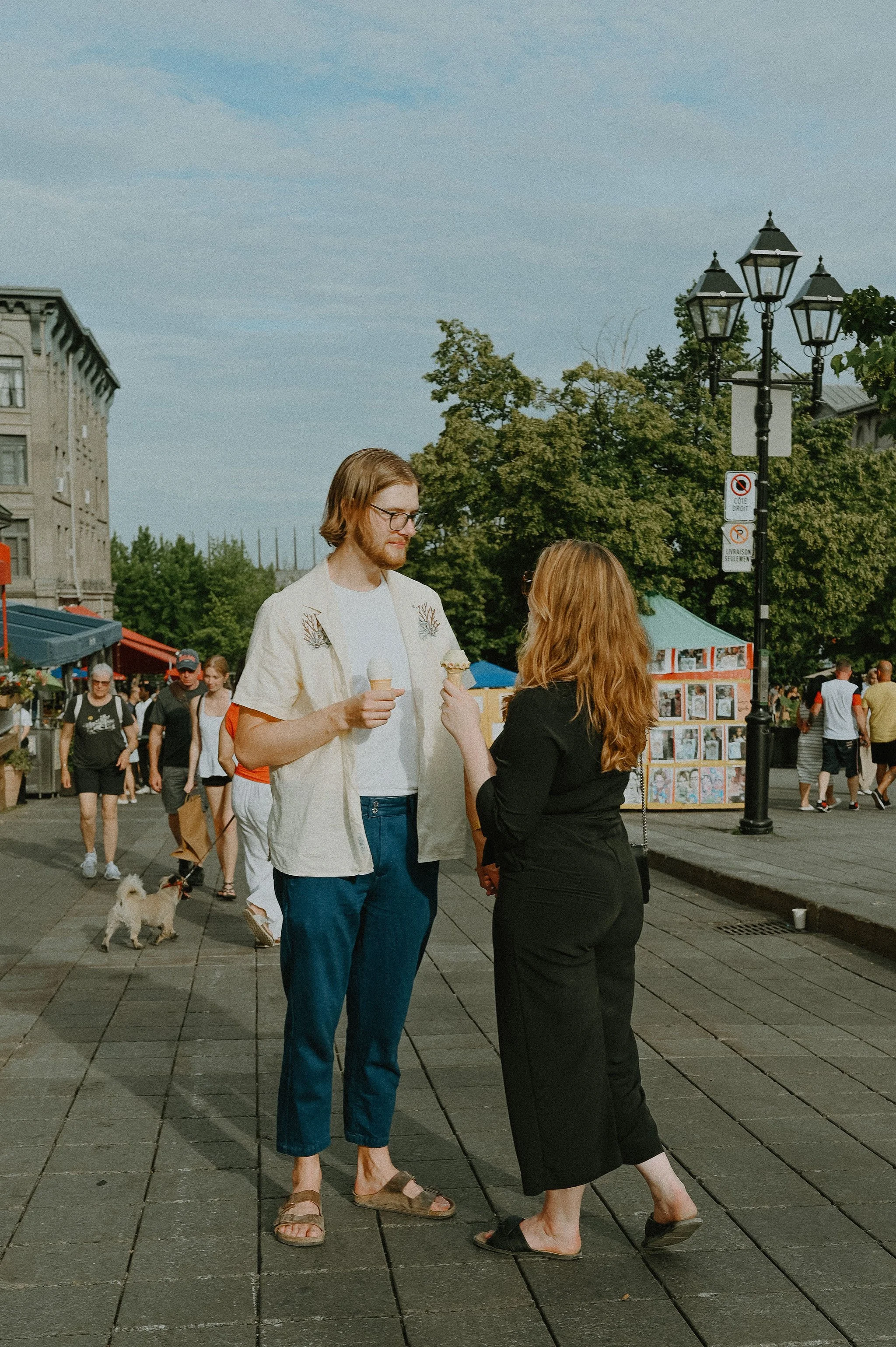 A man and woman talking on a sidewalk at an outdoor market, holding ice cream cones. The woman is wearing black, and the man is wearing a light-colored shirt. There are other people and market stalls in the background.