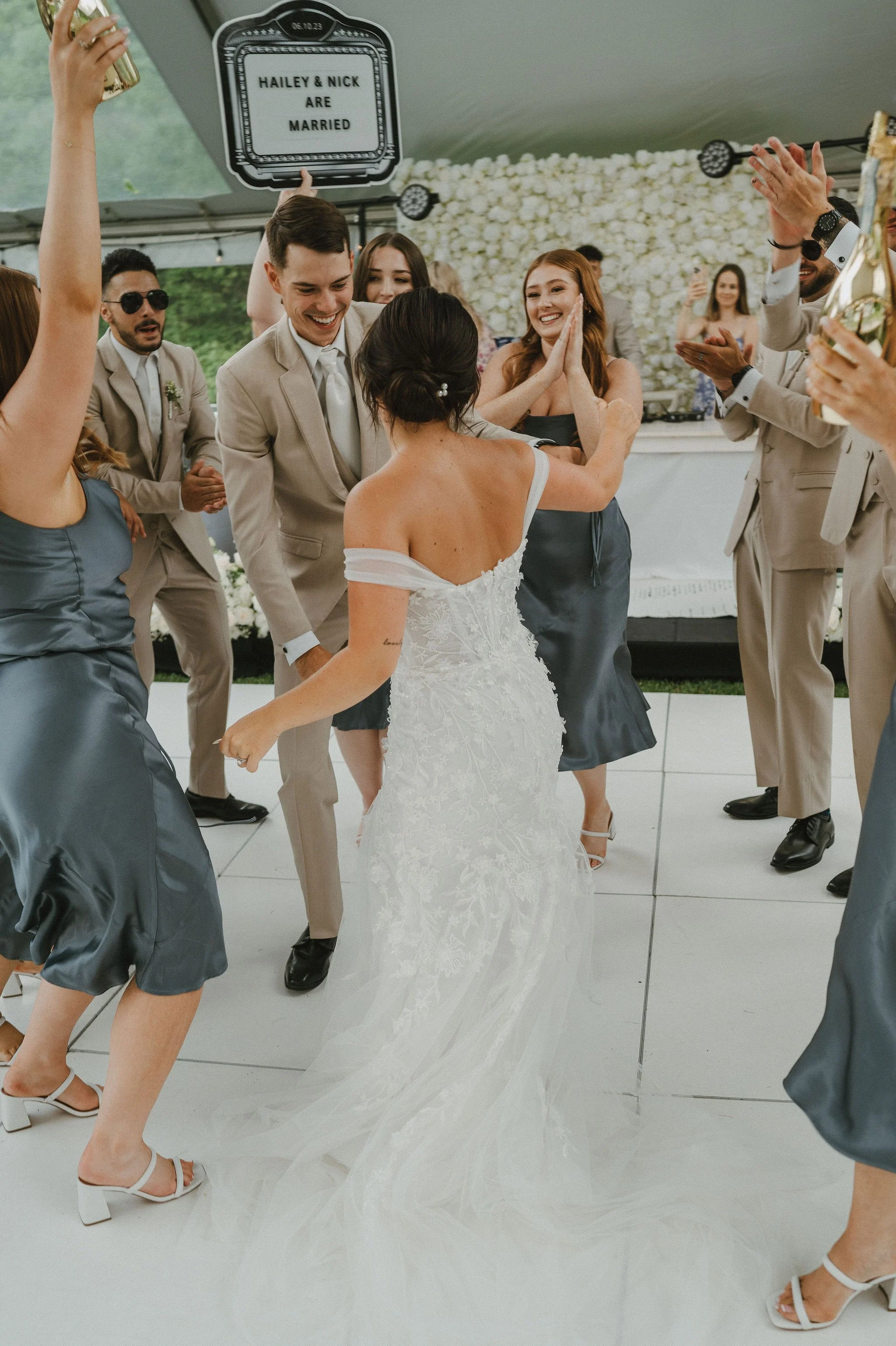 Wedding celebration with the bride in a white gown dancing with guests under a tent with a sign that reads 'Hailey & Nick Are Married' in the background.