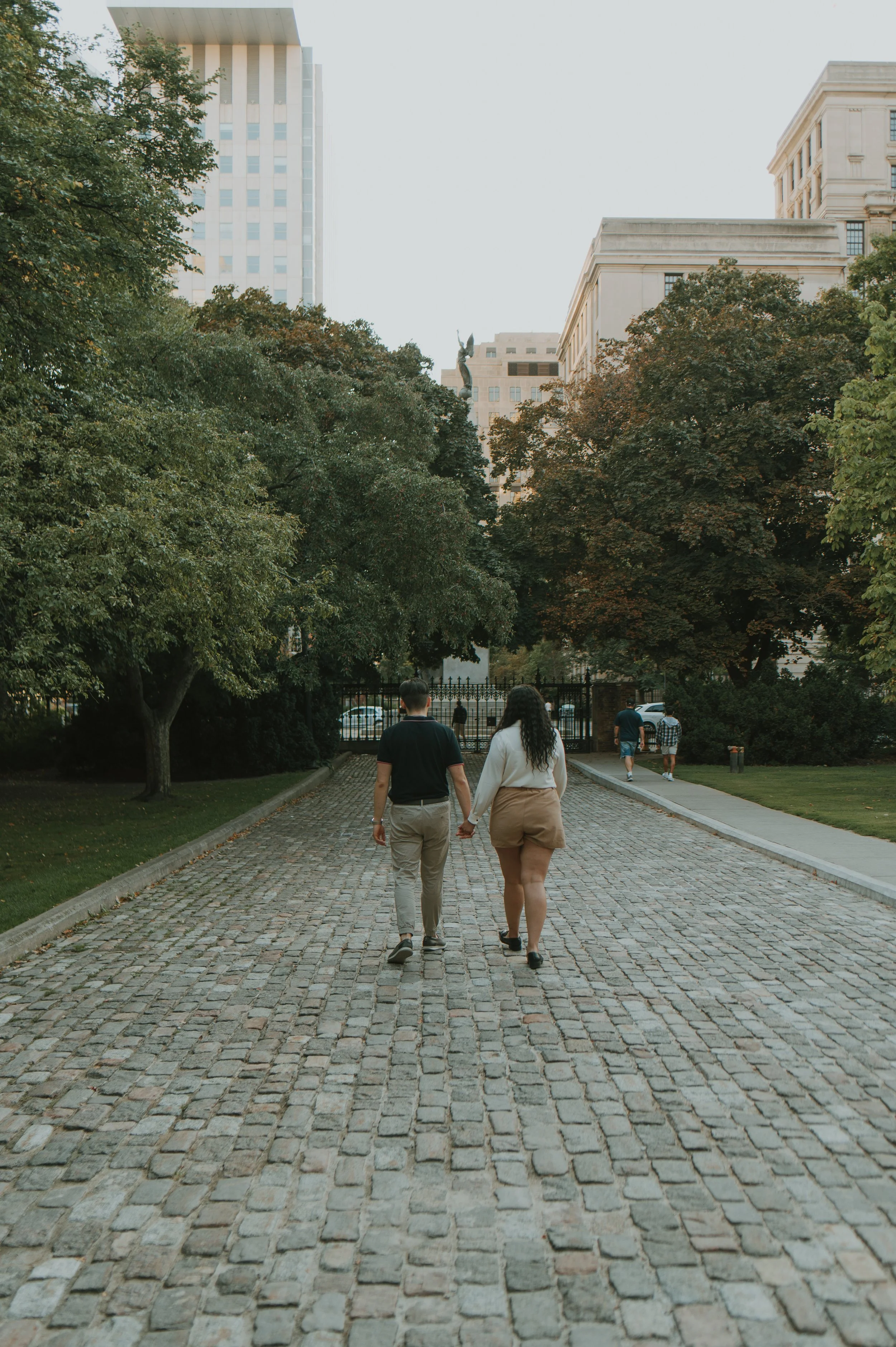 A couple walking hand in hand on a cobblestone path in a city park surrounded by trees, with tall buildings in the background.