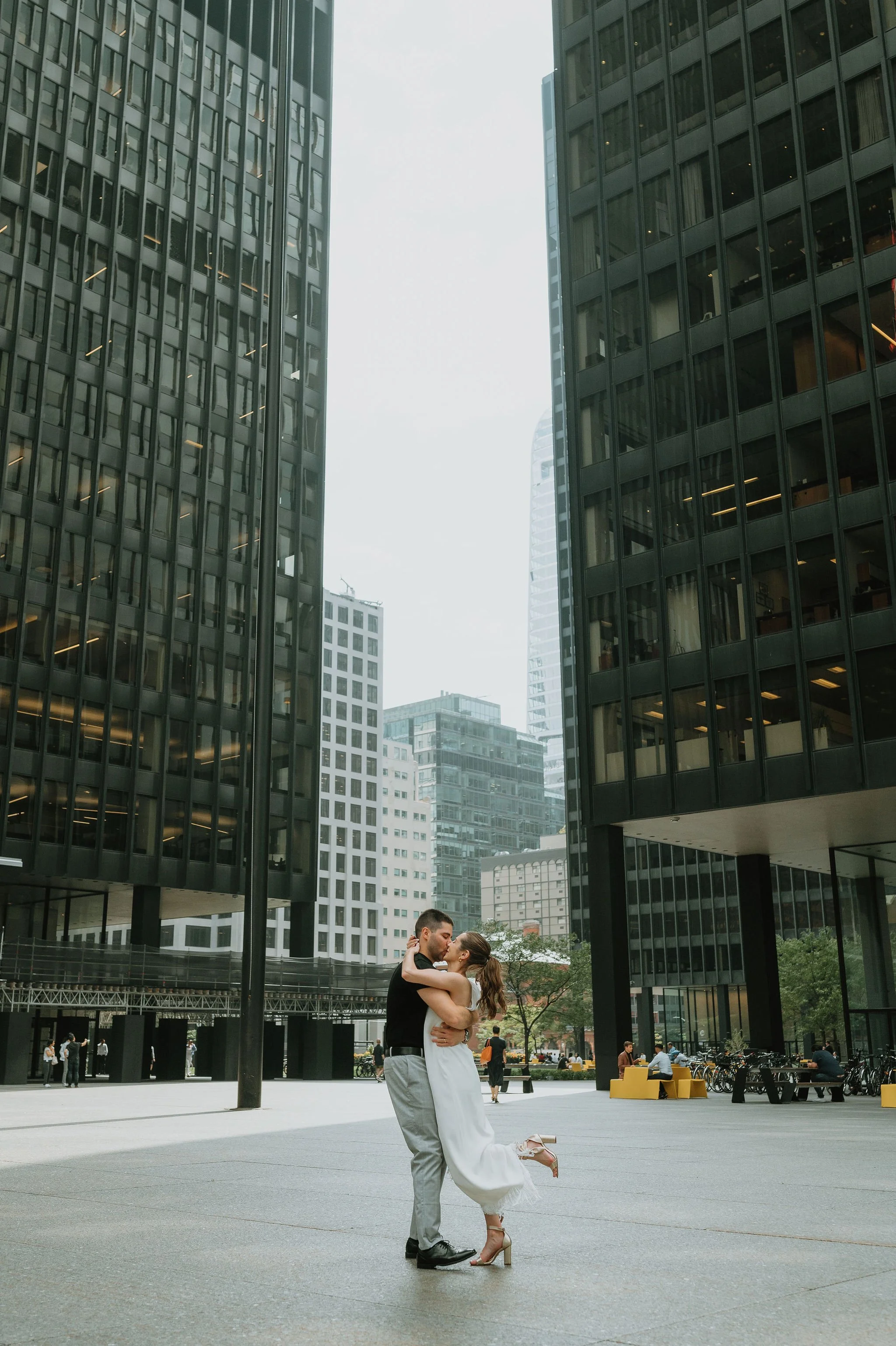 A romantic couple, embracing and about to kiss, dancing in an open urban plaza surrounded by tall modern office buildings during daytime.