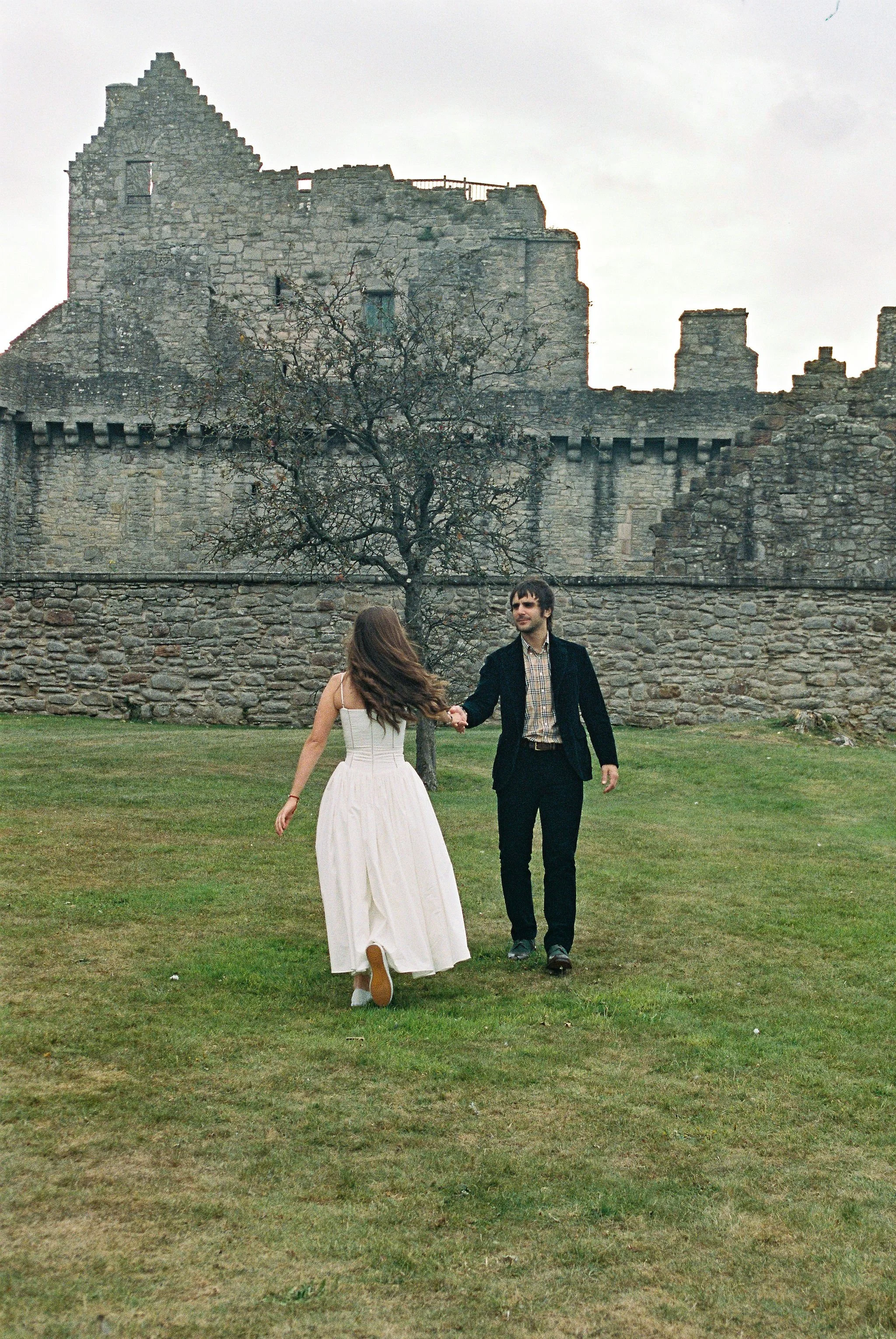 A man and woman holding hands and walking on grass in front of an old stone castle, with the woman wearing a white dress and the man in a dark suit.