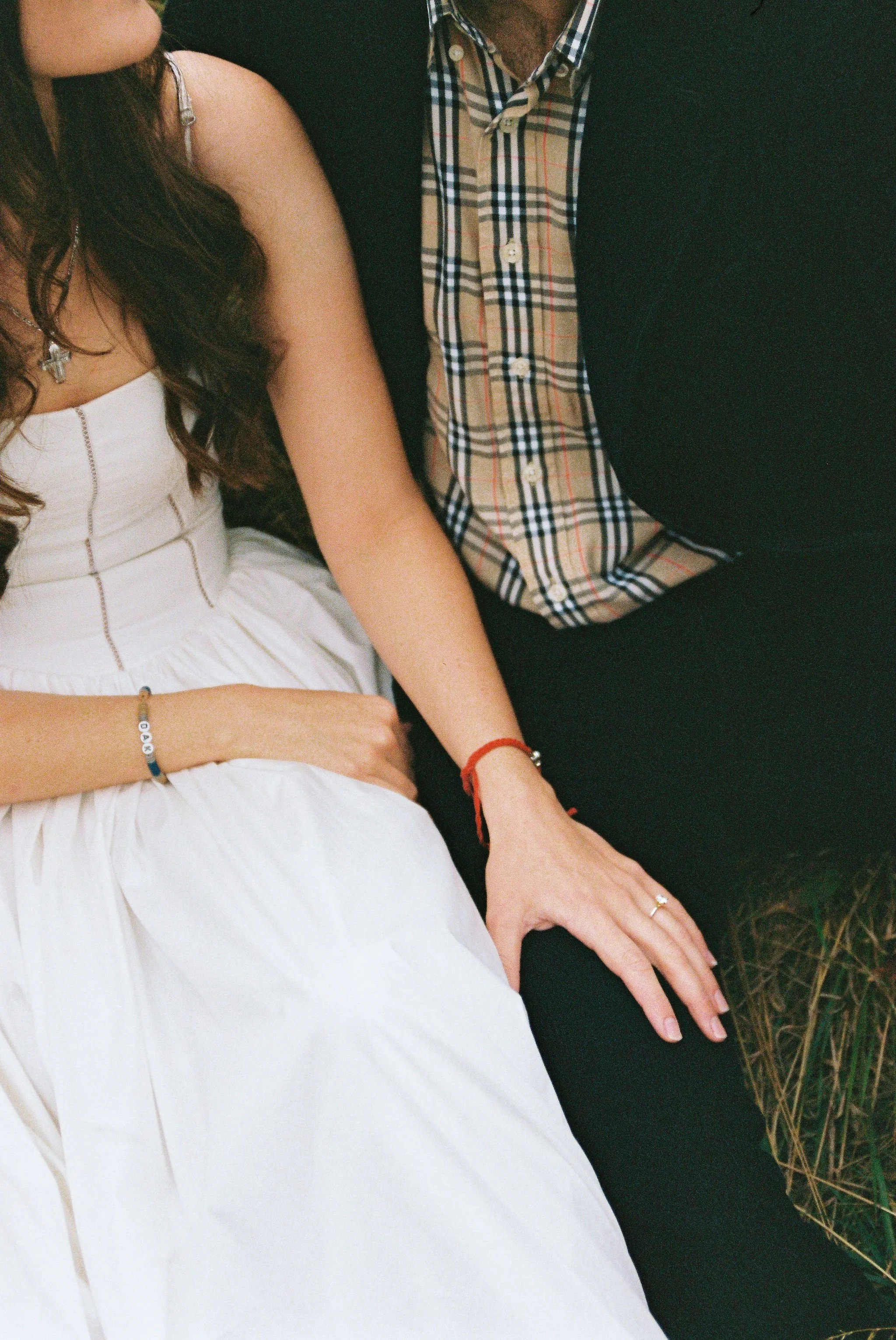 Close-up of a woman in a white dress and a man in a checkered shirt and black blazer sitting outdoors. Their hands are resting near each other.