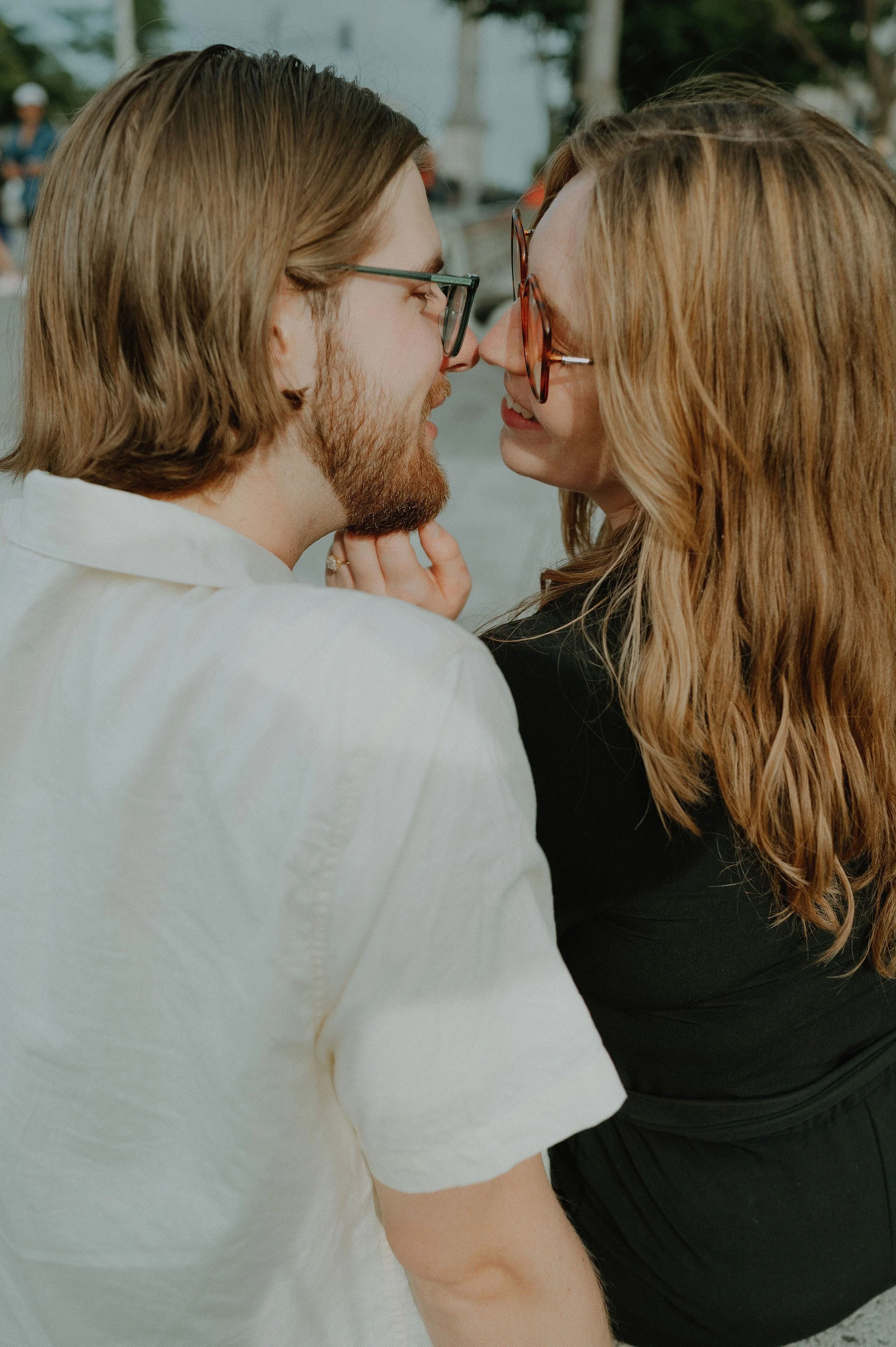 A couple with long hair and glasses face each other closely, touching noses, smiling, outdoors.