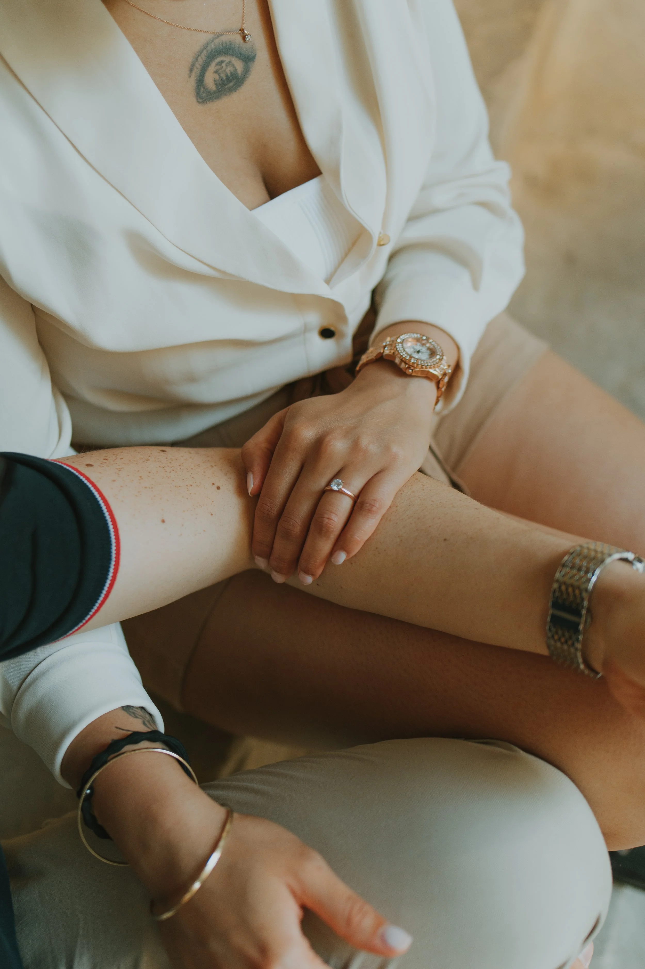 Close-up of two women's hands resting on a person's leg, with one hand wearing a ring and the other wearing a watch. The women are dressed casually, with one wearing a white blouse and the other wearing a black shirt. The setting is indoor with a neu