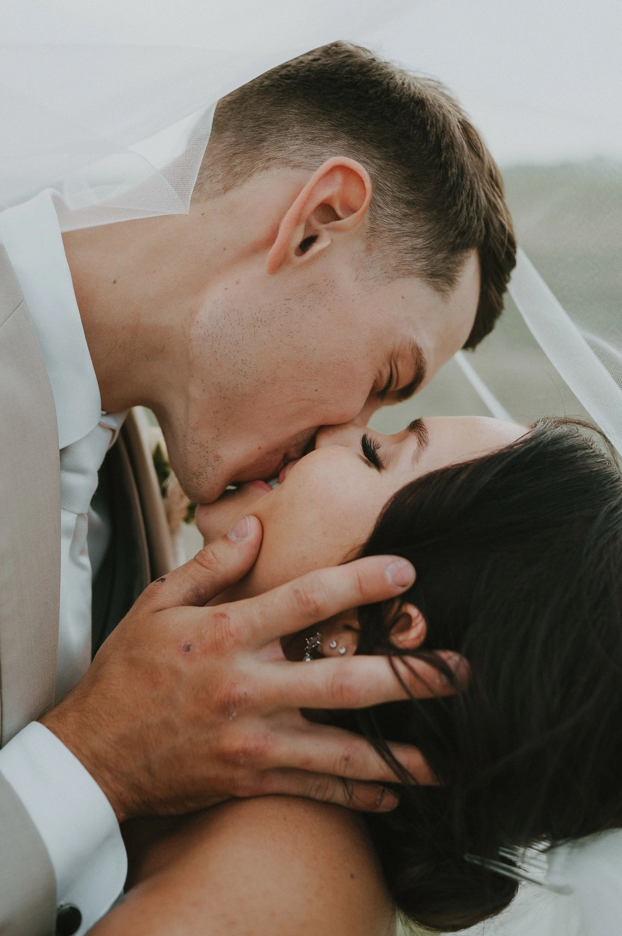 A close-up of a newlywed couple sharing a kiss with the groom gently holding the bride's face, under a white veil on a wedding day.