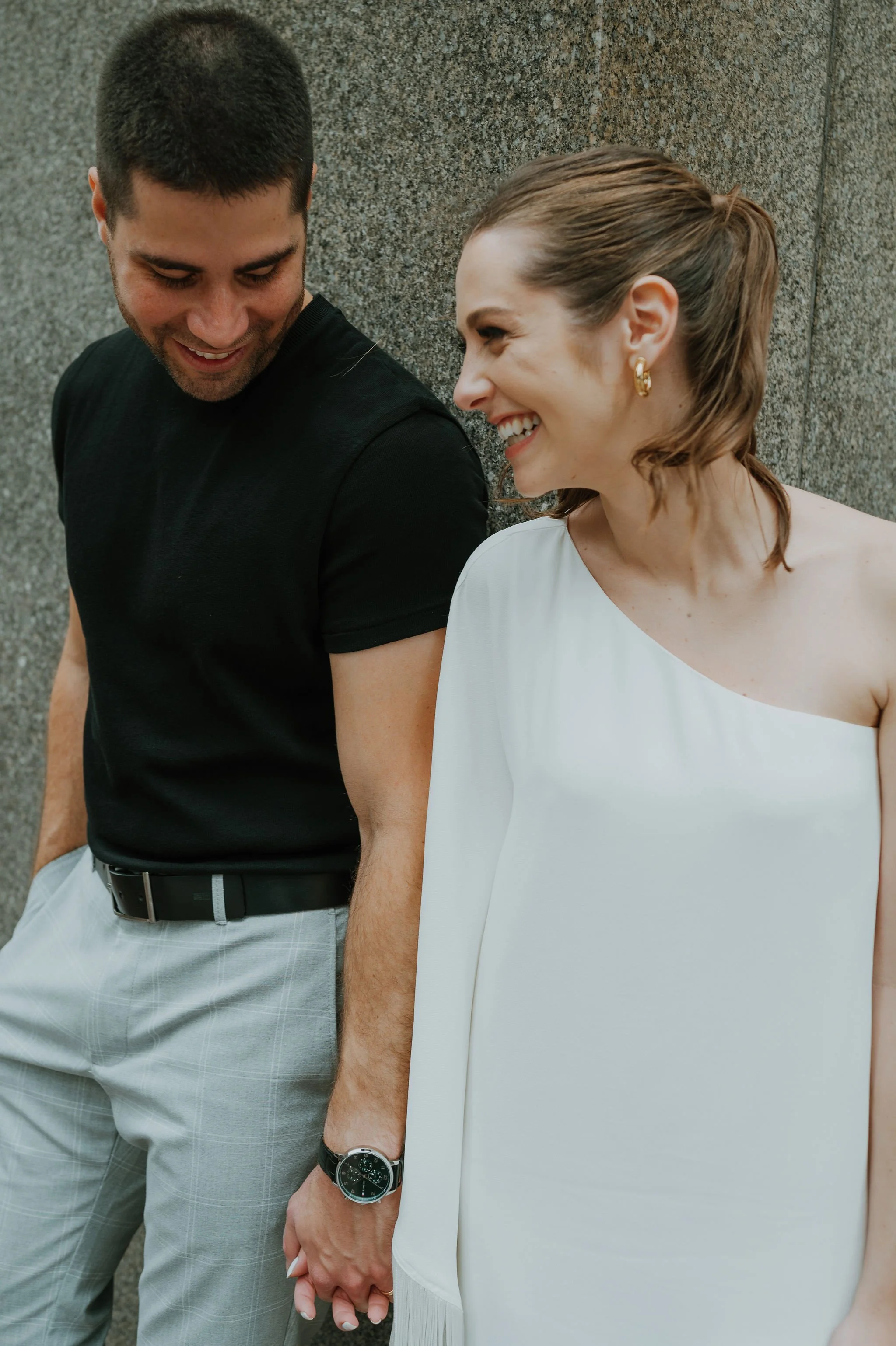 A man and woman smiling at each other, holding hands, standing against a stone wall. The woman is wearing a white dress with one shoulder, and the man is in a black shirt and light gray pants.
