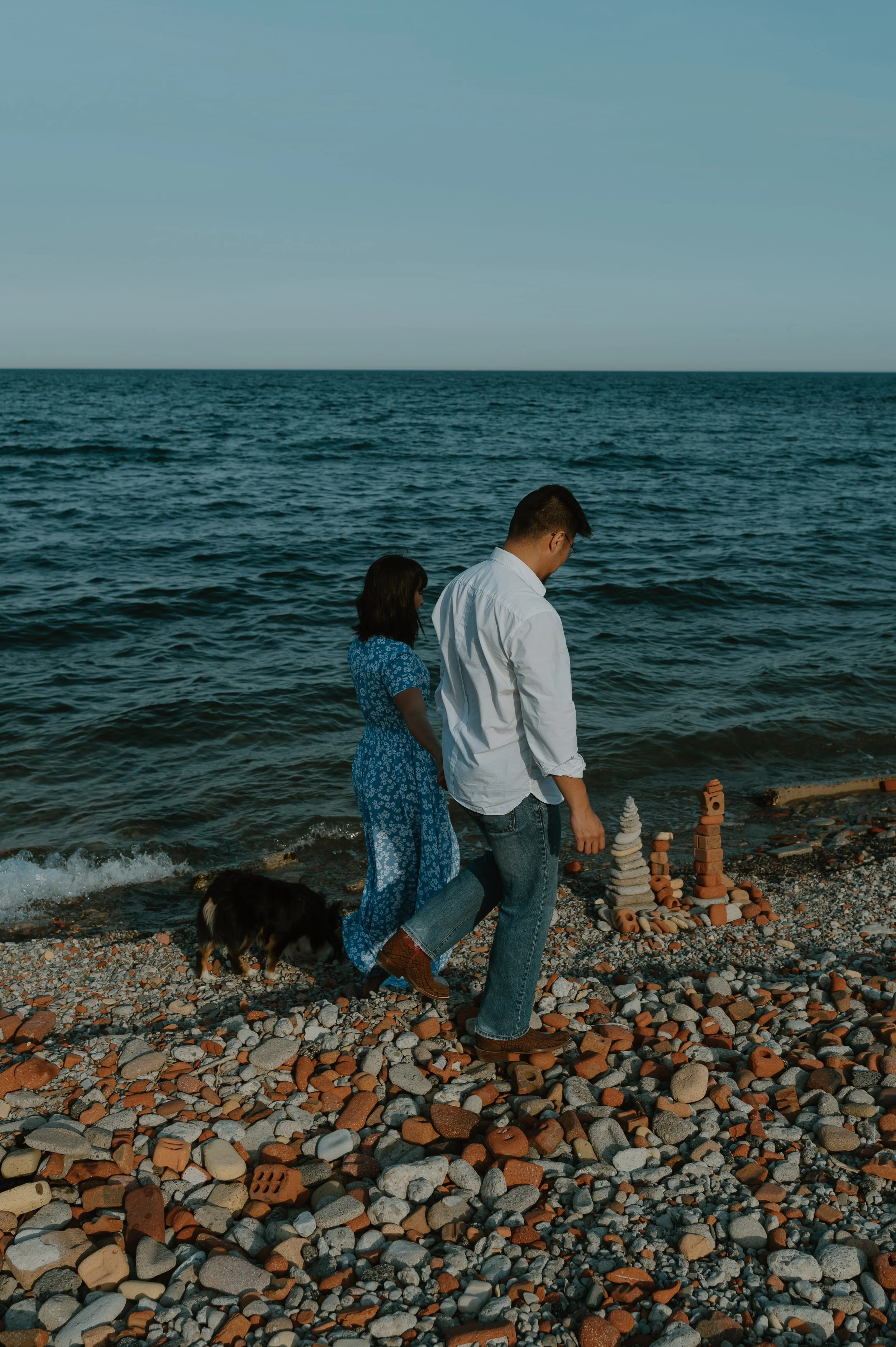 A man and a woman walking on a rocky beach with a dog, looking at stacked rocks near the water's edge, with the ocean and sky in the background.