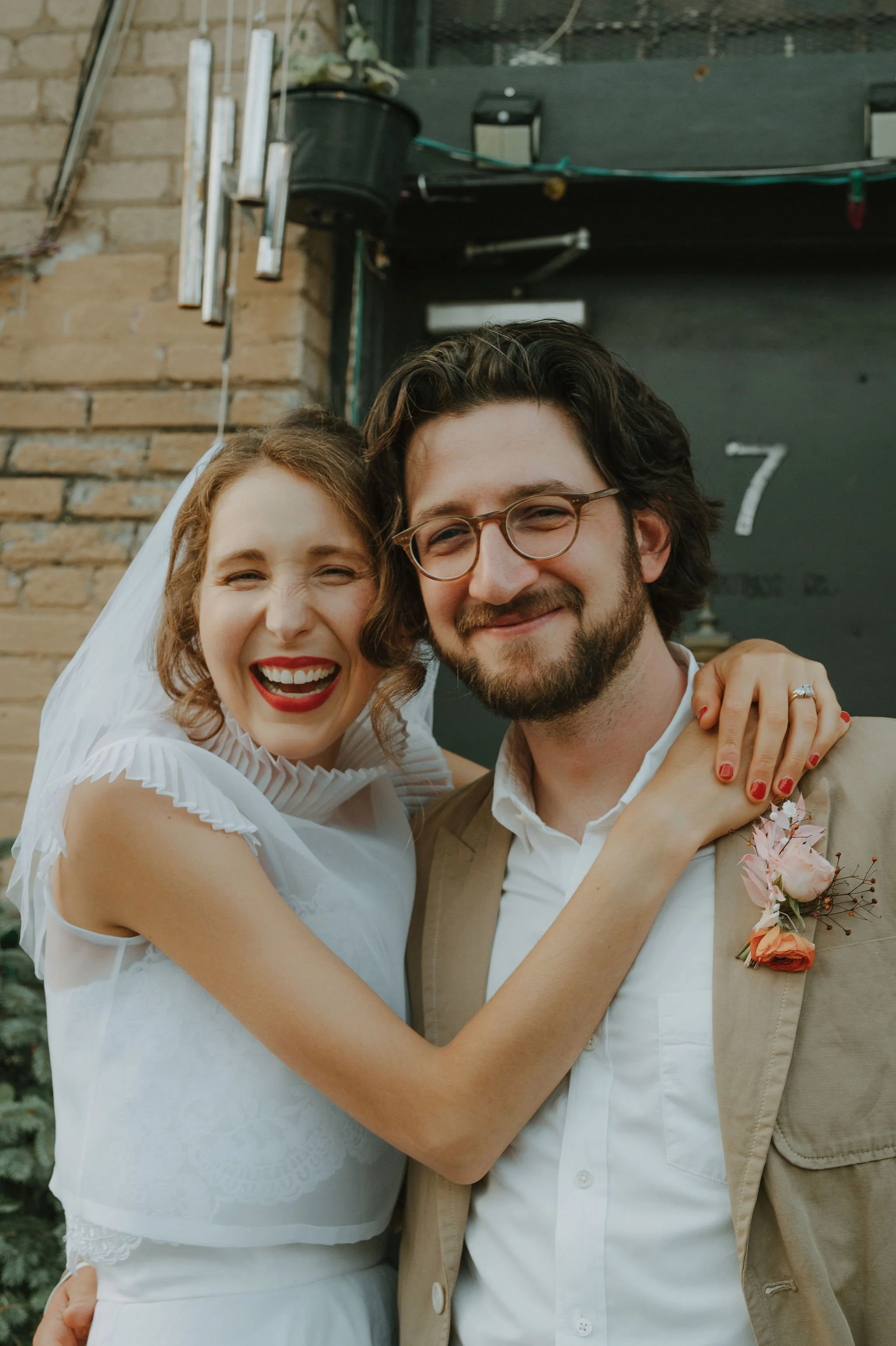 A happy bride and groom smiling together outdoors, with the bride wearing a white dress and the groom in a beige suit with a boutonniere, standing in front of a brick wall and a black door.