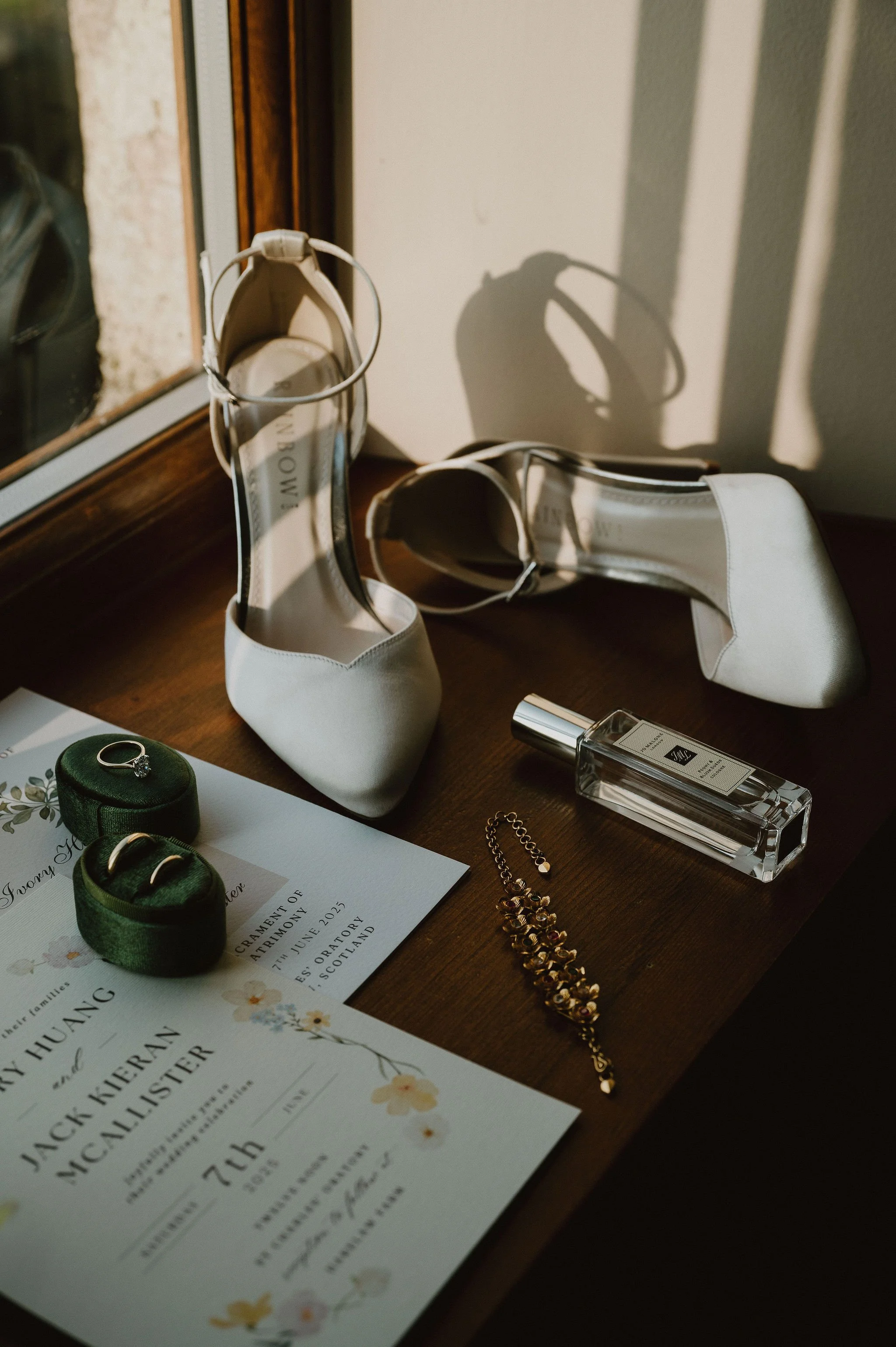White high heel shoes, a wedding invitation, rings on a green velvet ring box, earrings, a bracelet, and a perfume bottle on a wooden surface near a window.