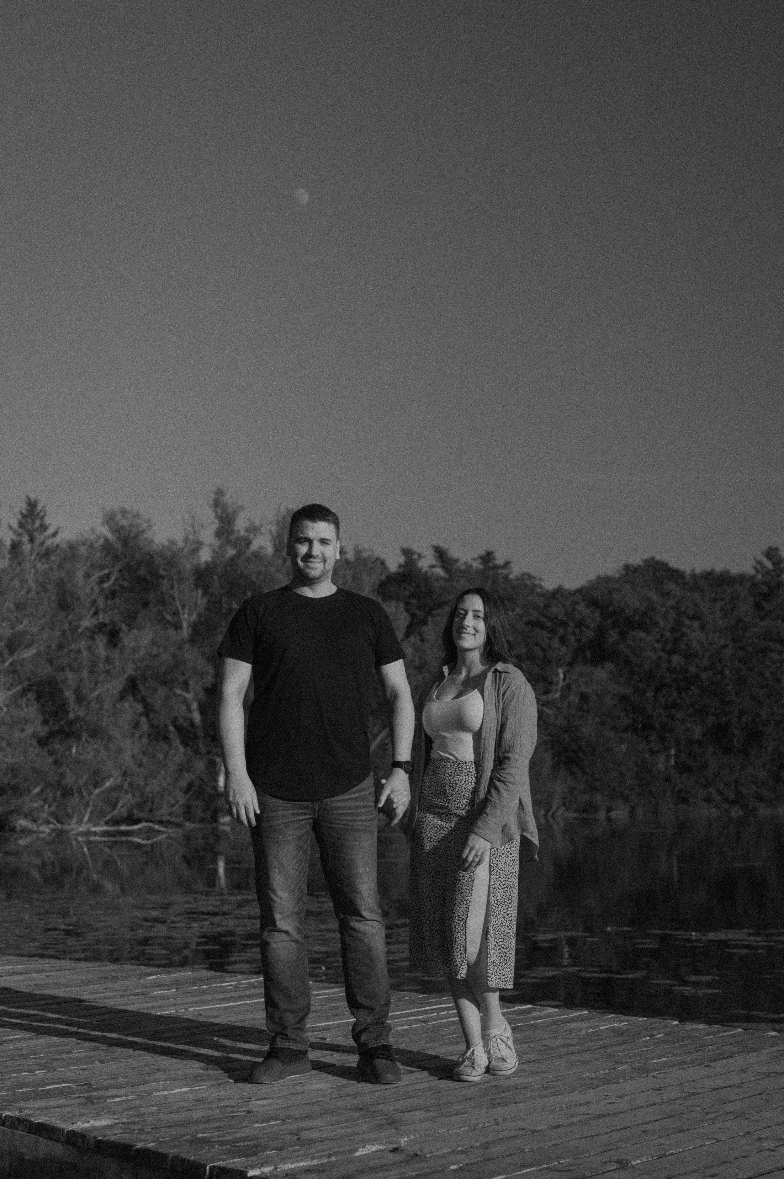 A black and white photo of a man and woman standing on a wooden dock outdoors with trees in the background and the moon visible in the sky.