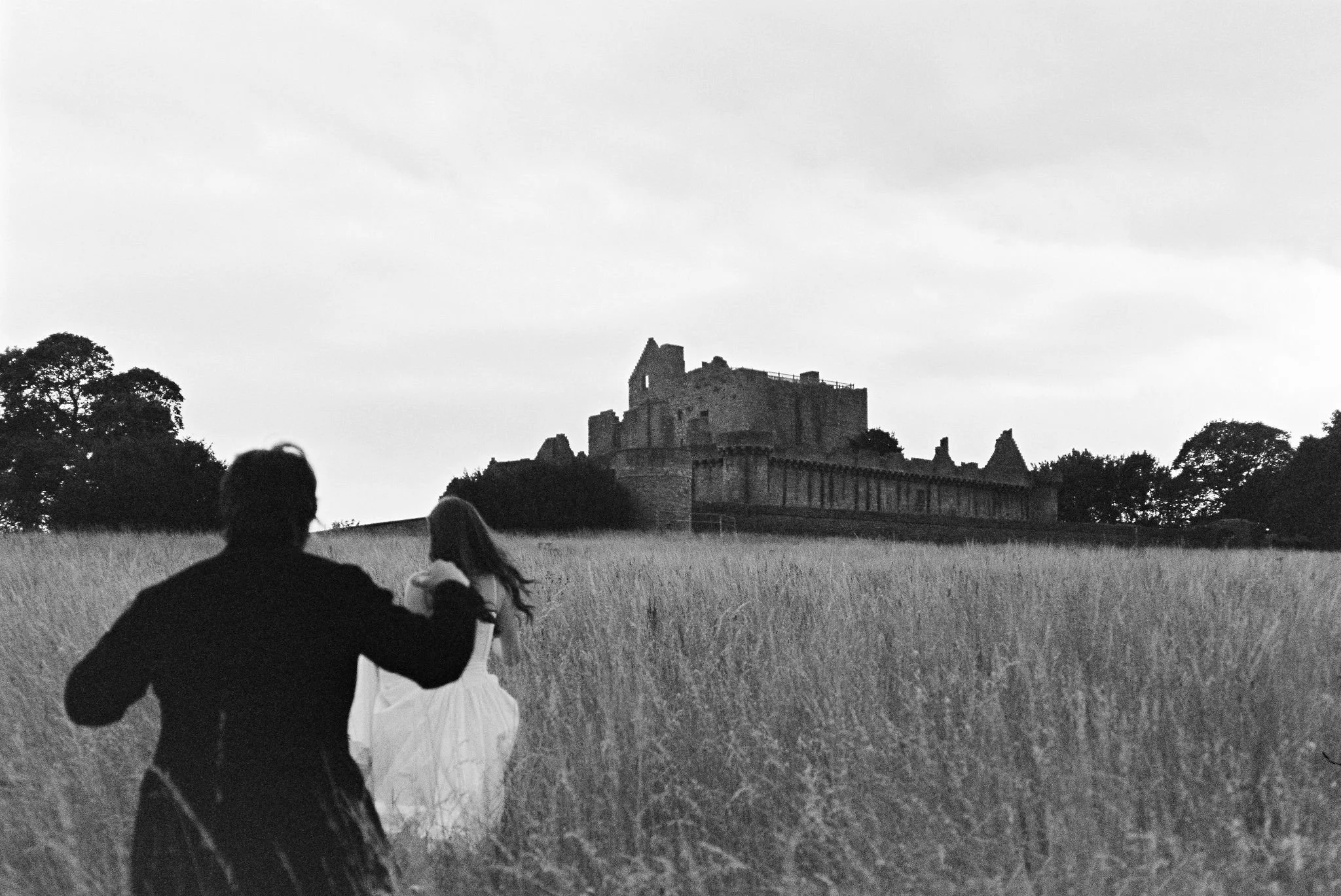 Silhouettes of a man and a woman holding hands walking through a grassy field towards a castle on a hill, in black and white.