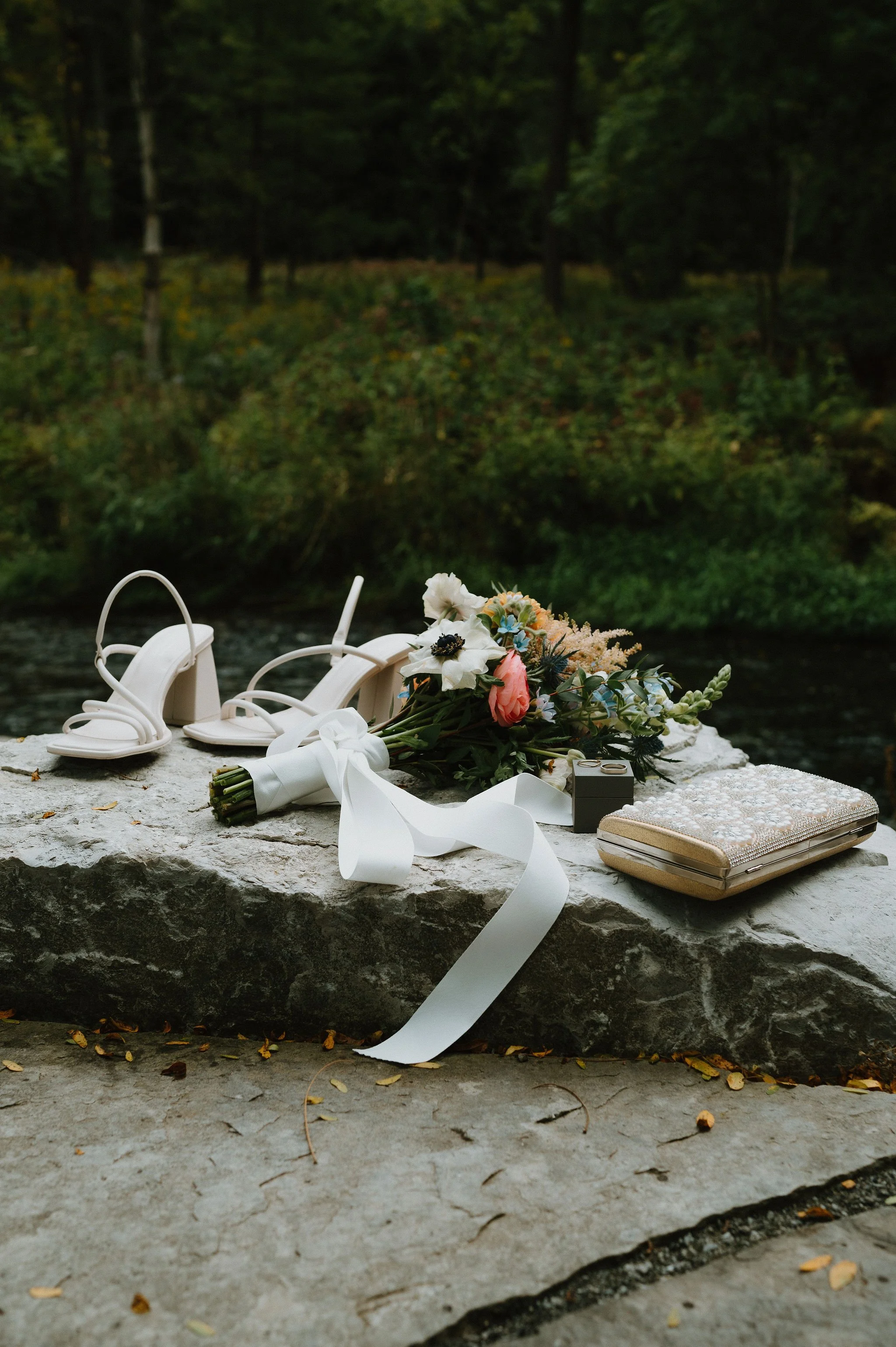 Wedding shoes, a bouquet of flowers, a small box, and a decorative clutch purse placed on a large rock outdoors with a wooded background.