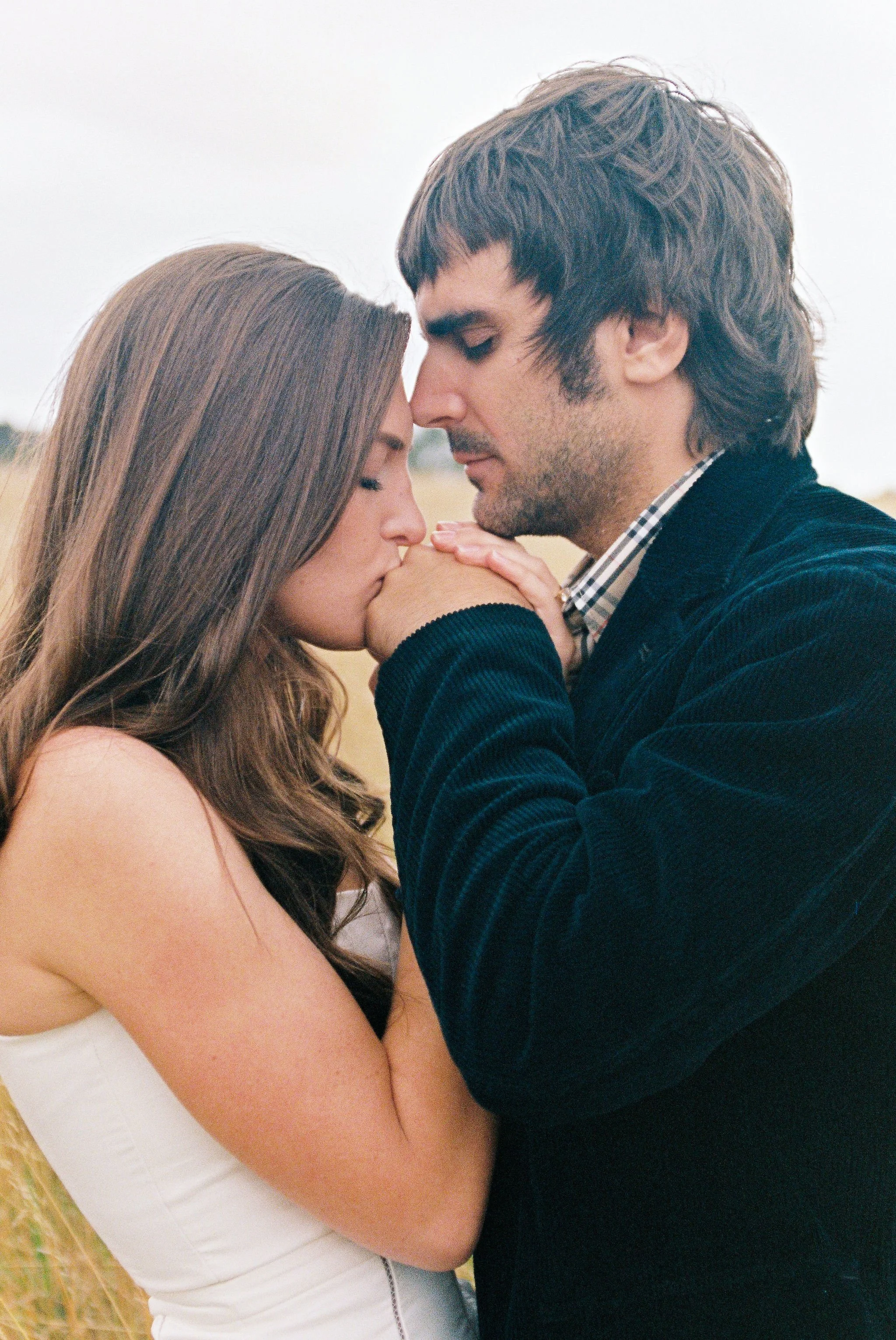 A couple sharing an intimate moment outdoors, with the woman kissing the man's hand and their foreheads touching.