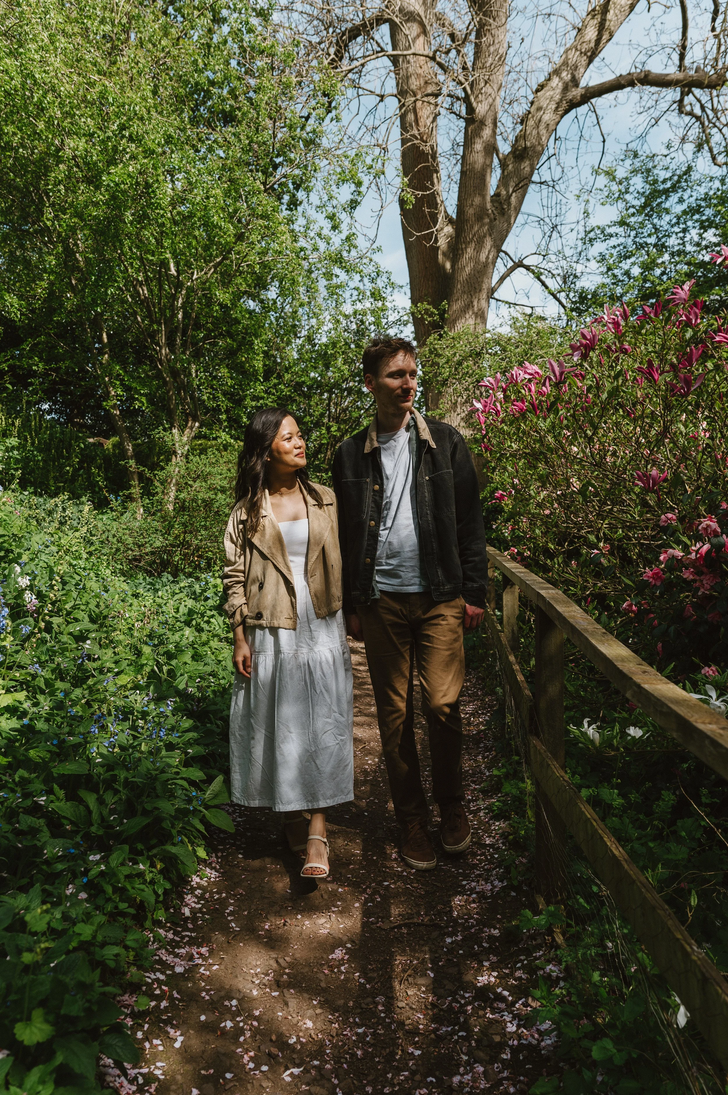 Two people, a woman in a white dress and beige jacket and a man in a gray shirt and brown pants, walk along a garden path surrounded by lush greenery and blooming pink flowers.