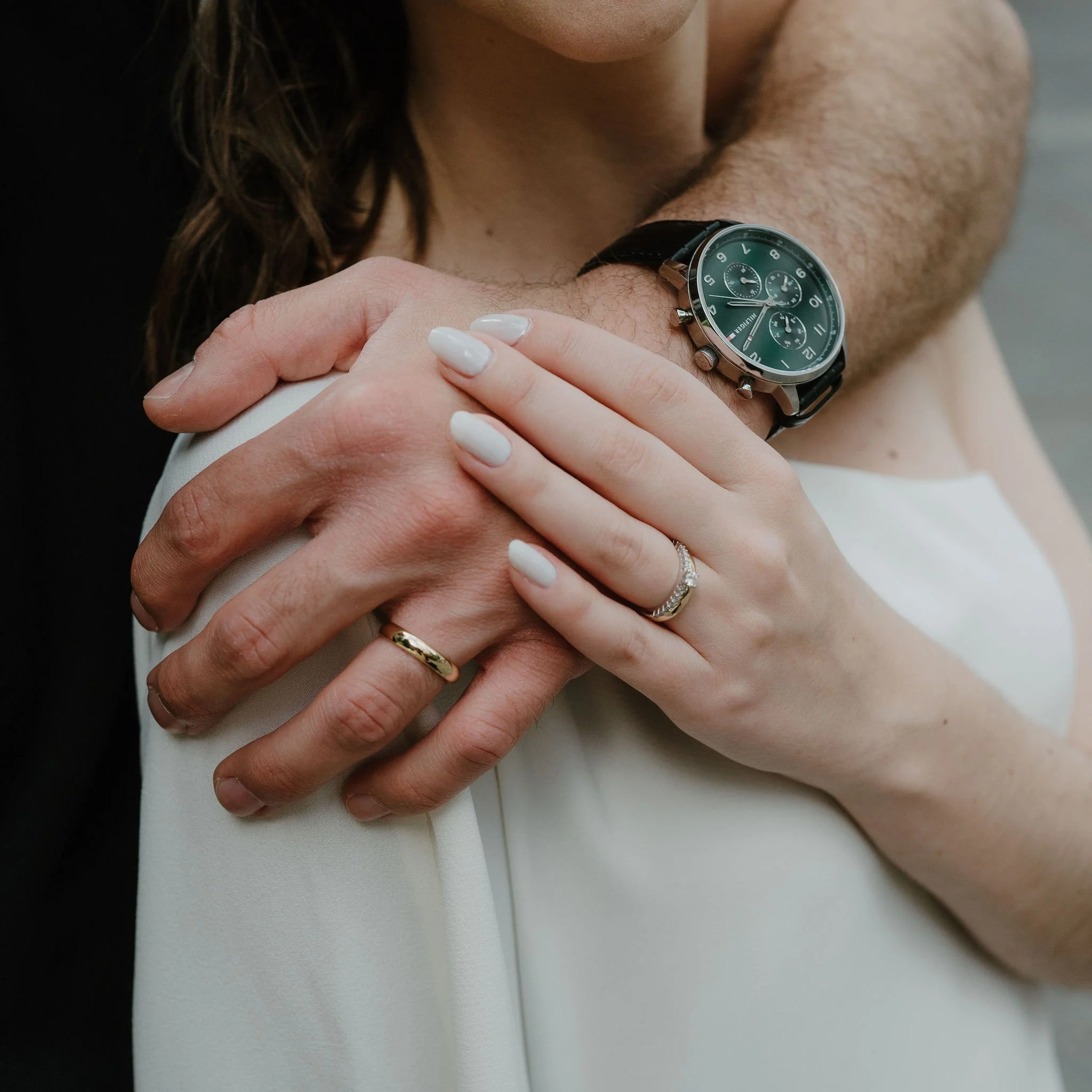 Close-up of a couple's hands, showing wedding bands, with the woman's hand resting on the man's shoulder. The woman wears a diamond ring on her ring finger, and the man wears a gold band. The man has a black watch with a green face.