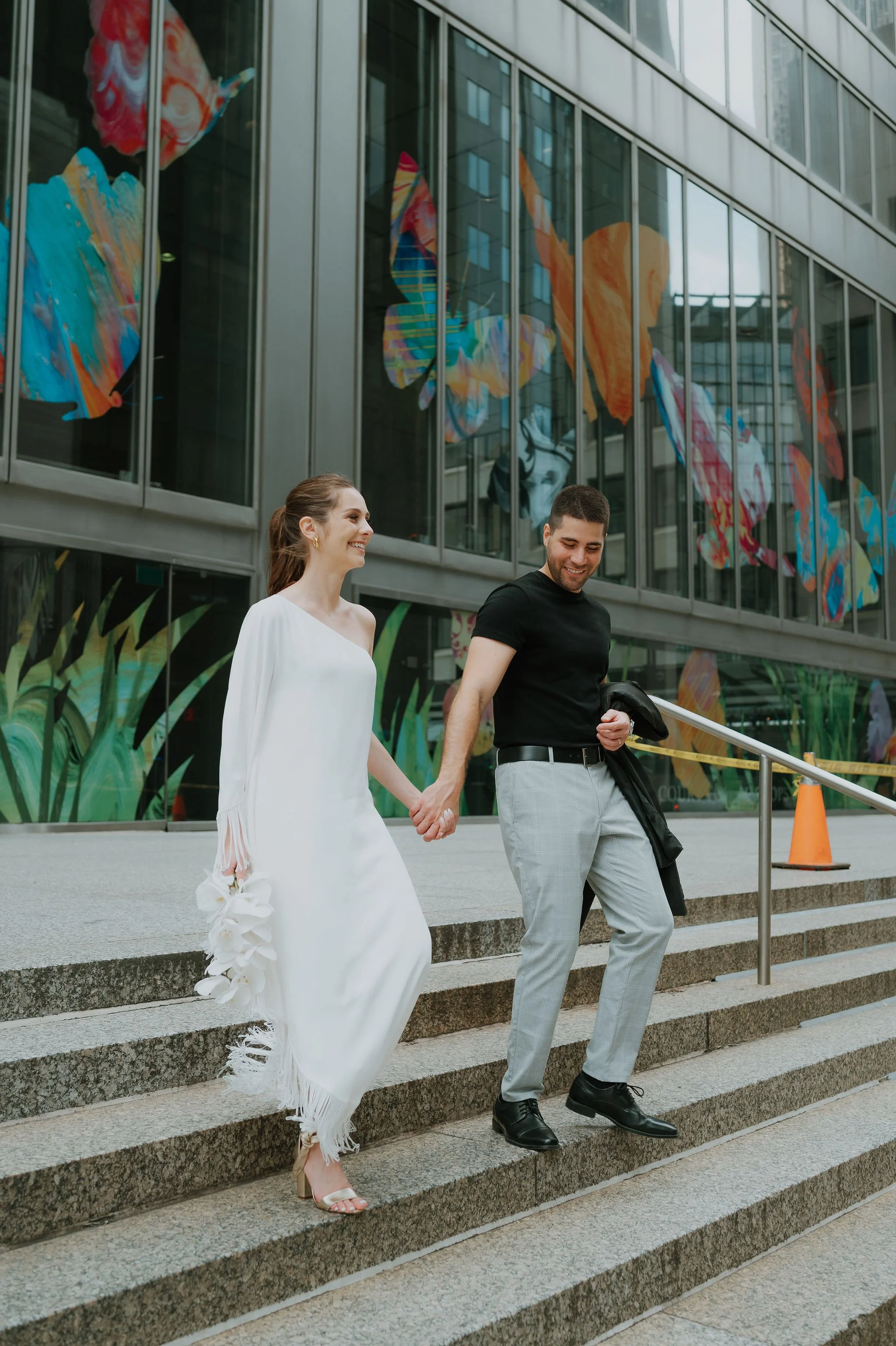 A happy couple holding hands and walking down outdoor stairs in front of a glass building with colorful butterfly and plant artwork.