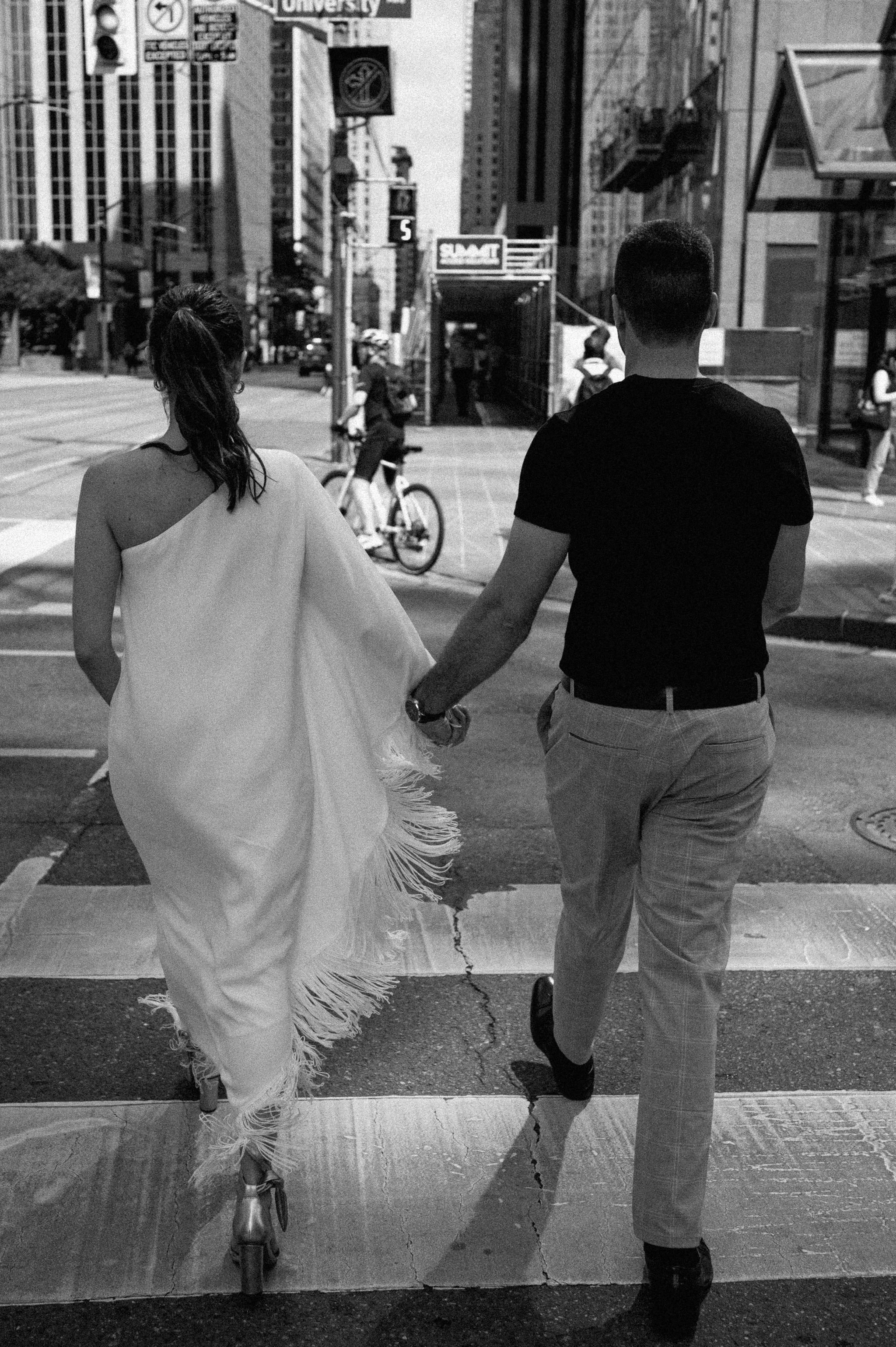 A black and white photo of a couple holding hands and crossing the street in a city with tall buildings and pedestrians.