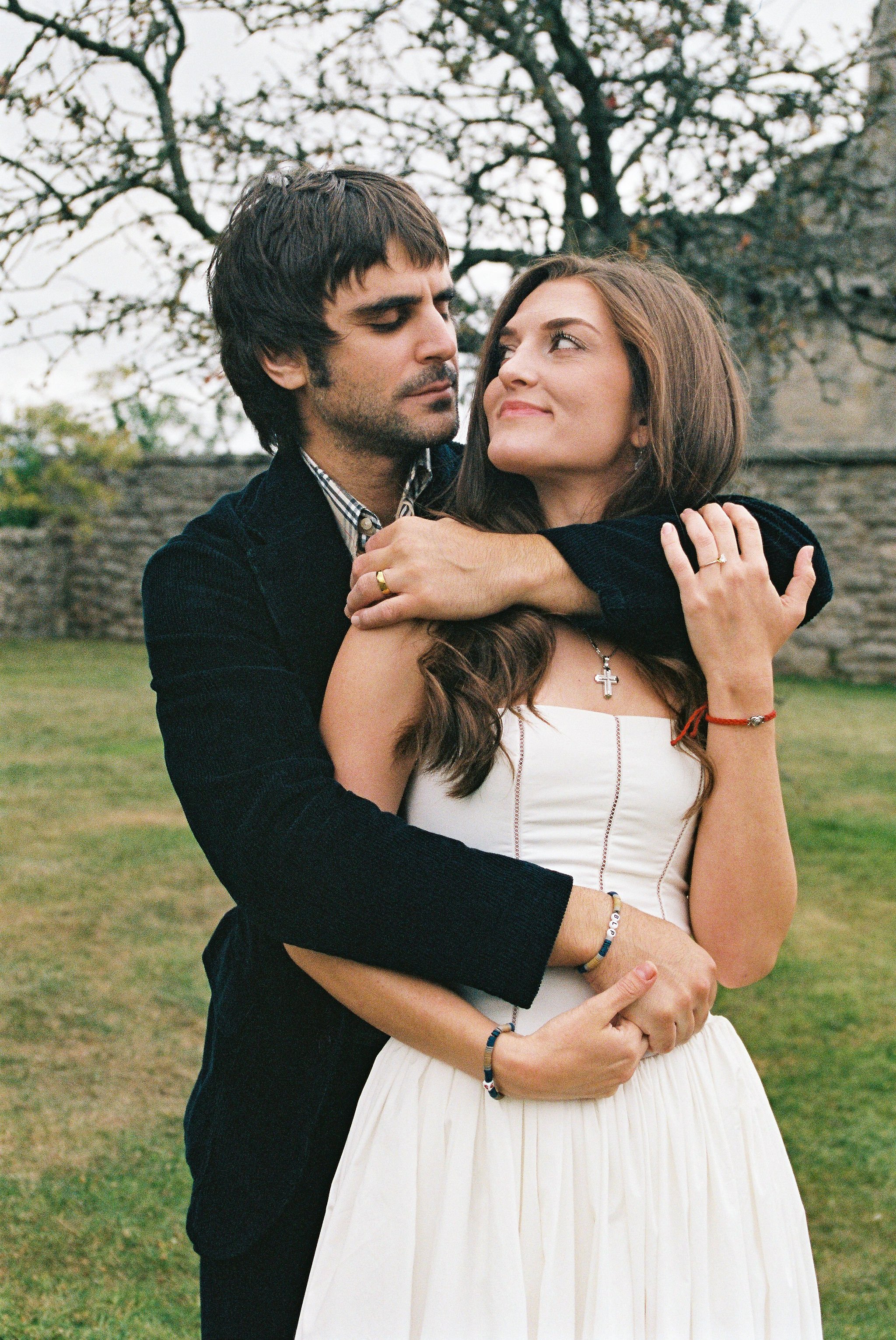A man hugging a woman in an outdoor setting with trees and a stone wall in the background.