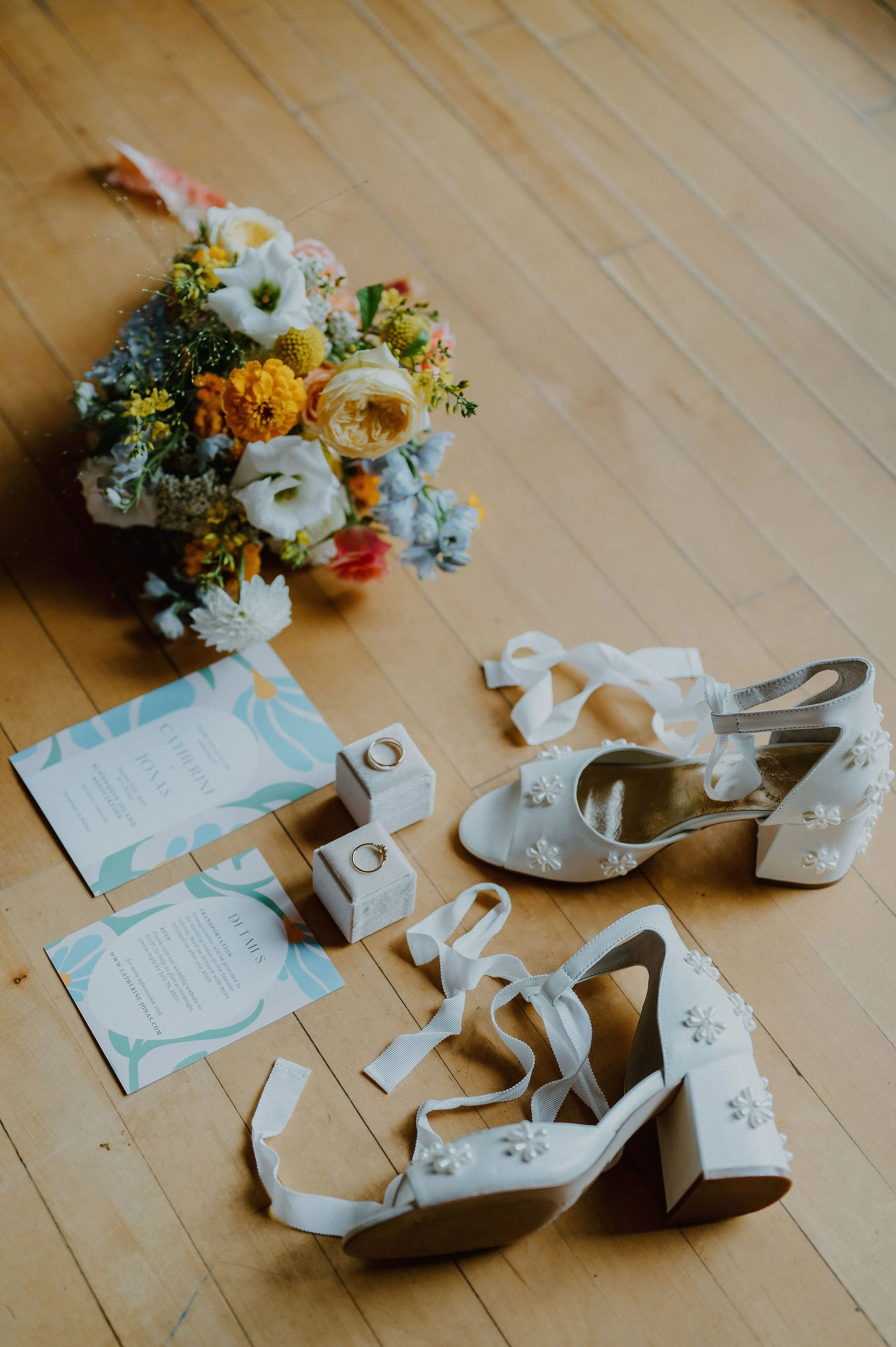A bridal bouquet with white, yellow, orange, and pink flowers, two wedding rings on white ring boxes, and a pair of white floral-embellished high-heeled shoes on a wooden floor.