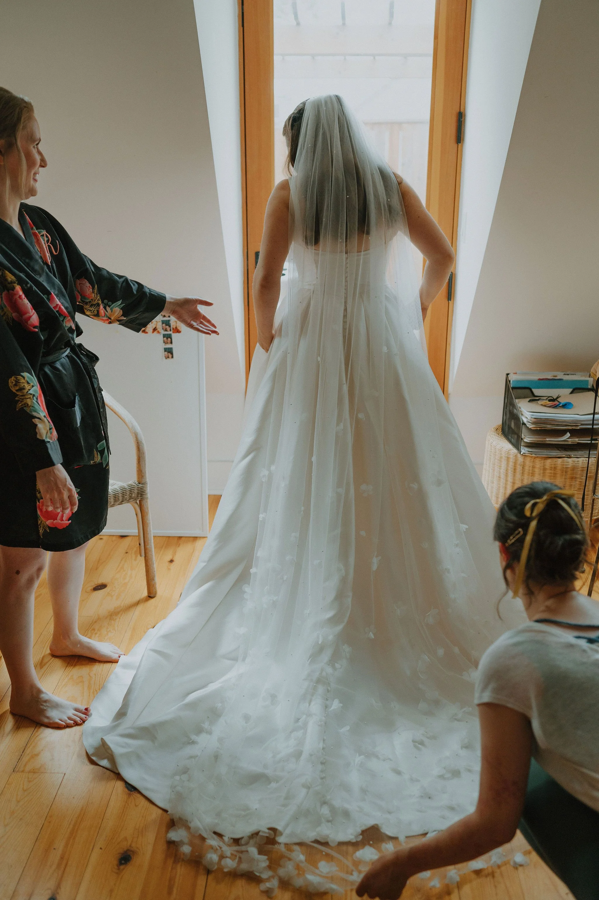 A bride in a white wedding dress and veil standing in front of a window, with two women helping her prepare, one woman gesturing towards her and another woman sitting nearby.