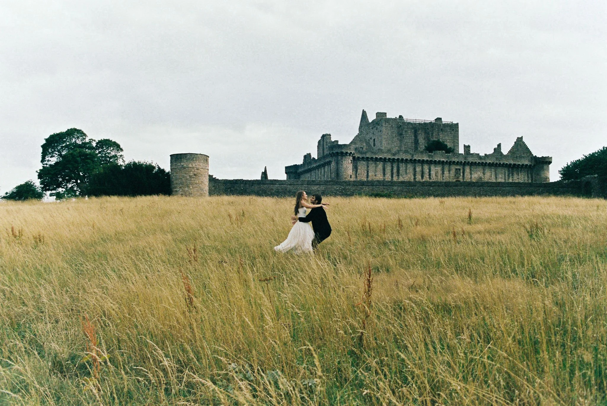 A couple dancing in a field of tall grass with a historic castle in the background.