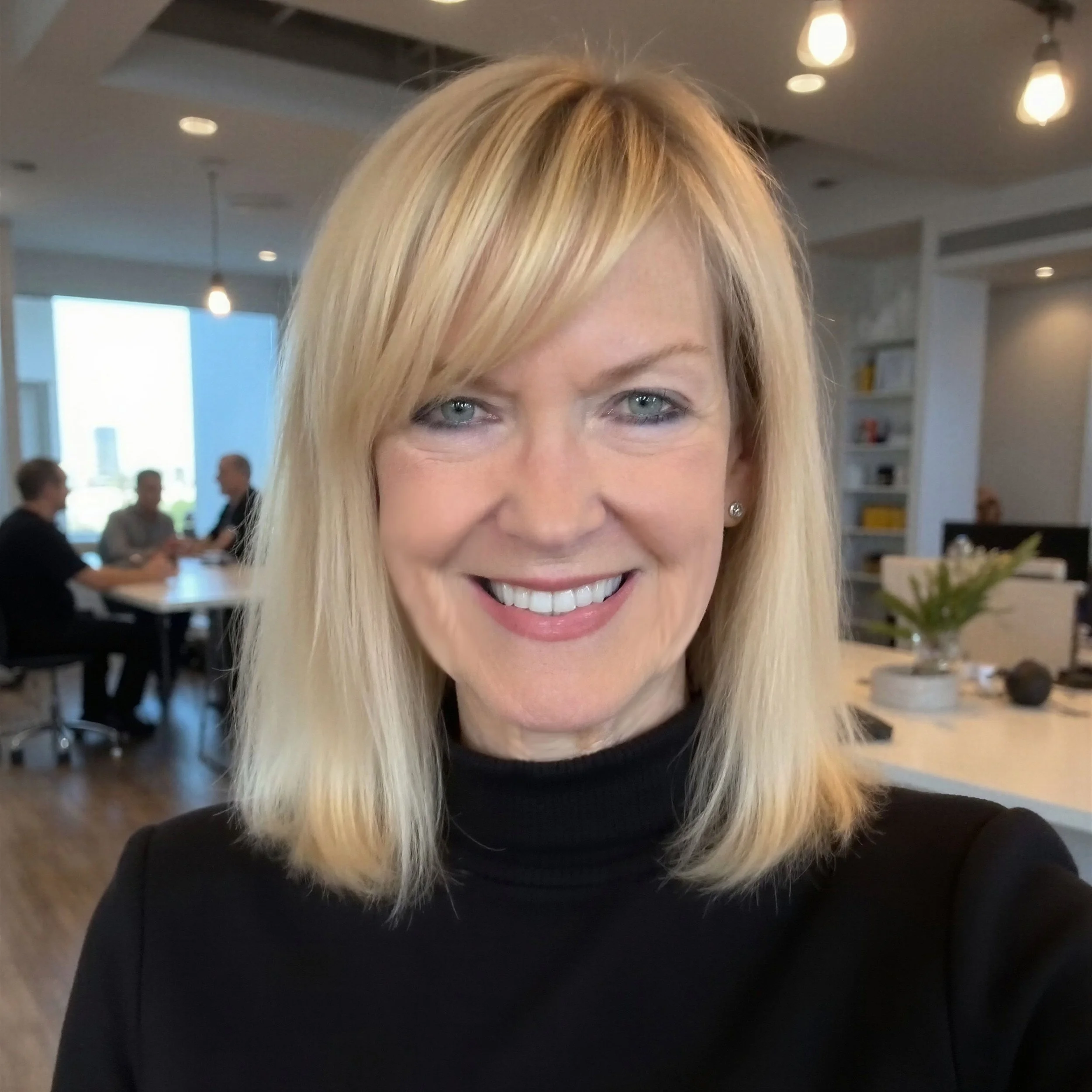 A smiling woman with blonde hair and blue eyes, wearing a black top, is in an indoor space with modern lighting and people in the background.