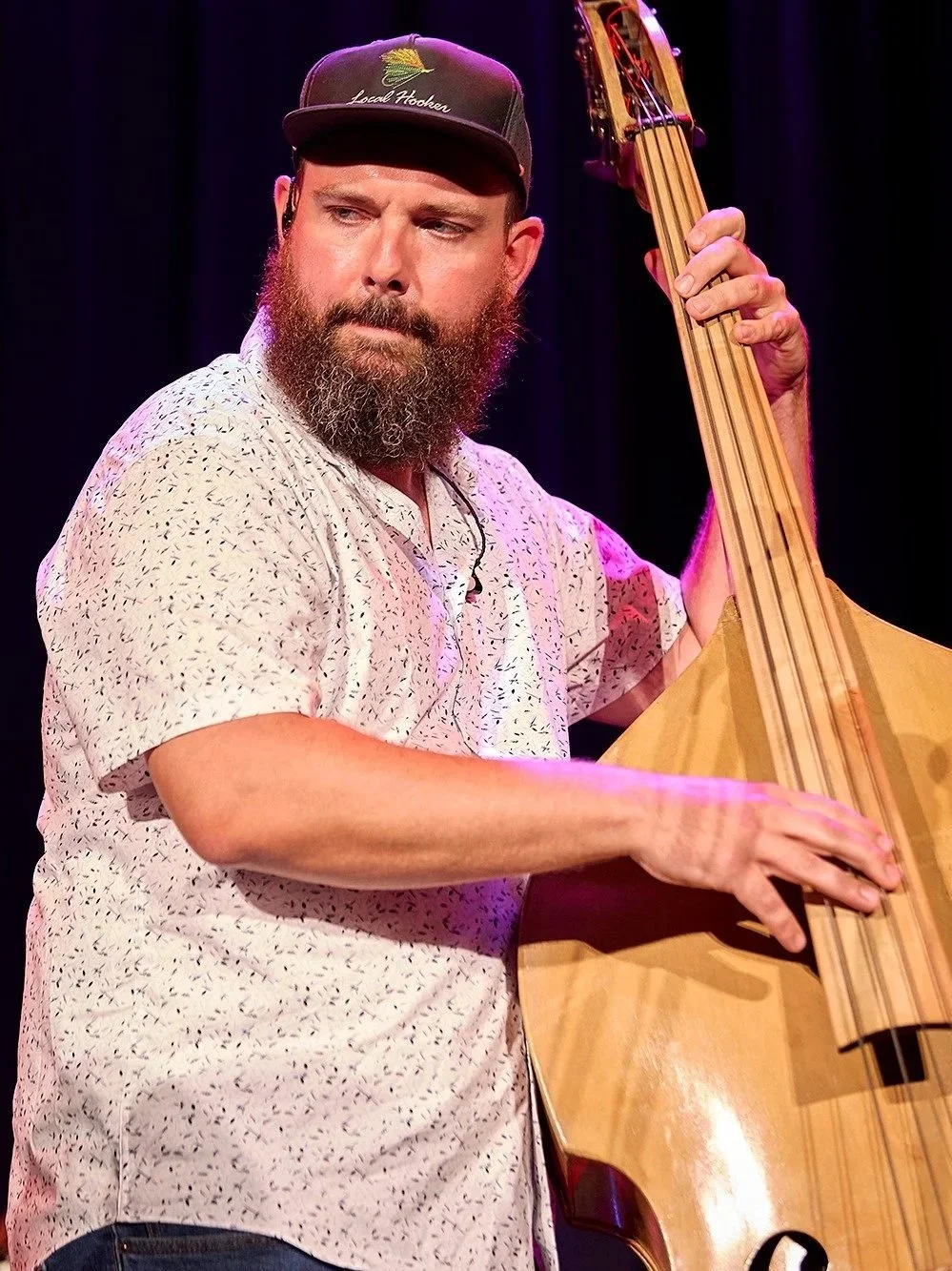 A man with a beard and baseball cap playing a double bass on stage.
