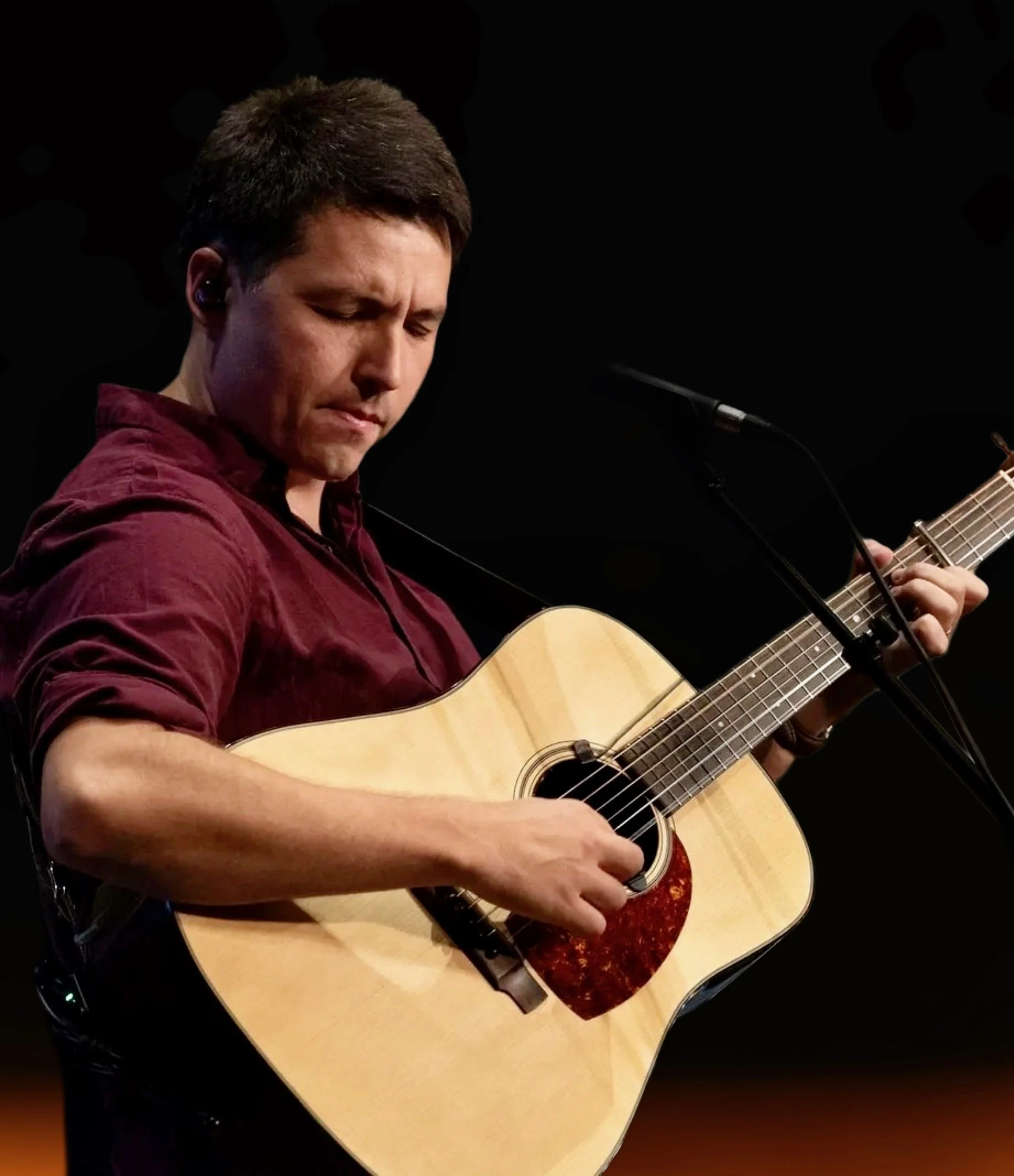 A man playing an acoustic guitar on stage, with eyes closed, wearing a red long-sleeve shirt, in front of a black background.