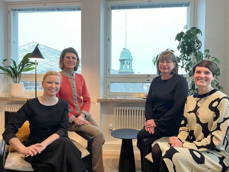 Neljä naista istuu tuoleilla hymyillen suoraan kameraan. Valoisassa taustassa näkyy viherkasveja sekä ikkunoita, joista avautuu näkymä kaupungin talojen katoille ja taivaalle.