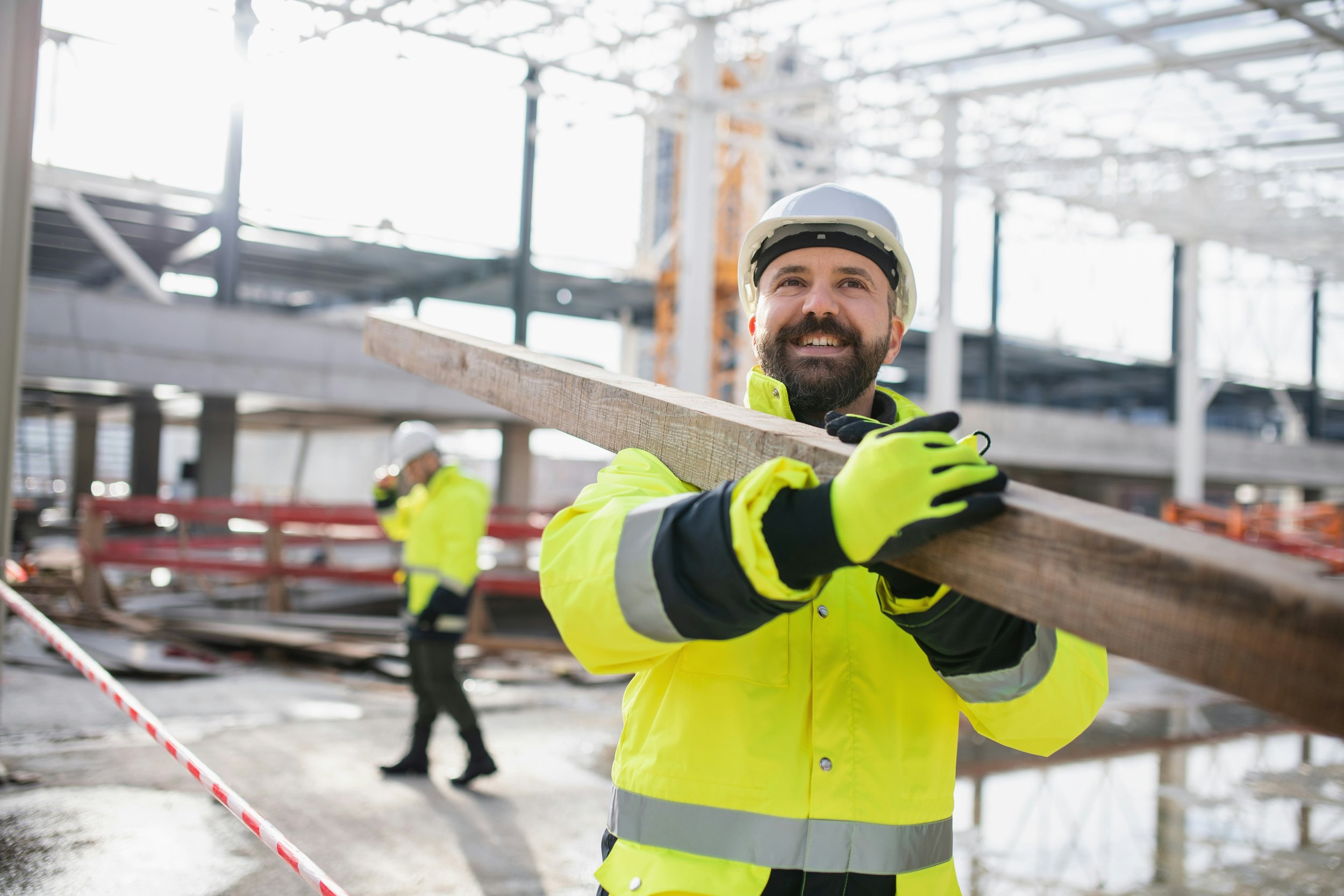 Iloinen rakennusalan työntekijä työvarusteissaan kantamassa lankkua.