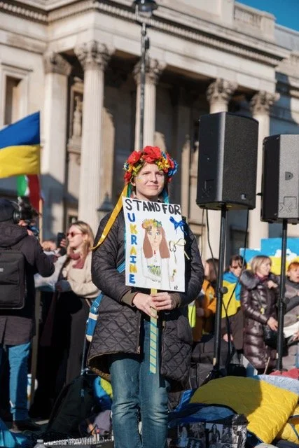 Henkilö seisoo ulkona aurinkoisella kaupungilla ja pitelee käsissään kylttiä, jossa lukee "Stand for Ukraine". Kuva välittää vahvaa tukea ja solidaarisuutta Ukrainalle.