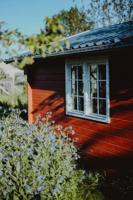 Kaunis punainen mökki, jossa valkokehyksiset ikkunat, peltikatto ja vieressä kukkaistutuksia.