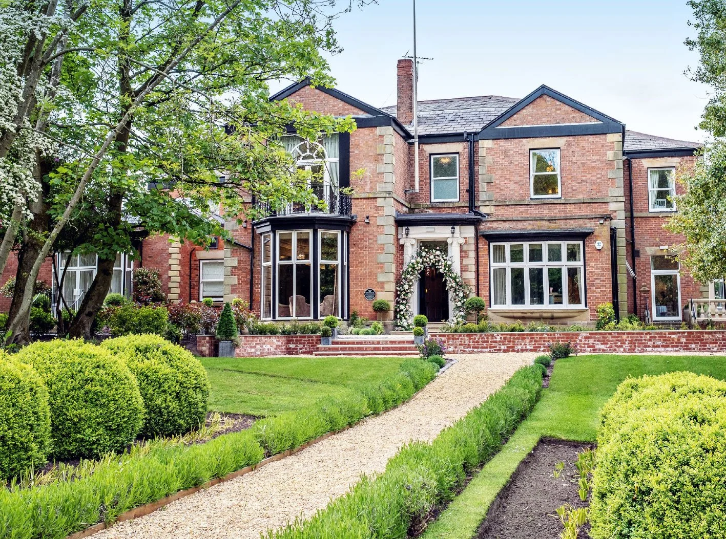 A large, elegant brick house with a manicured lawn and garden, decorated with flowers and bushes, featuring a central doorway with an arch of flowers.