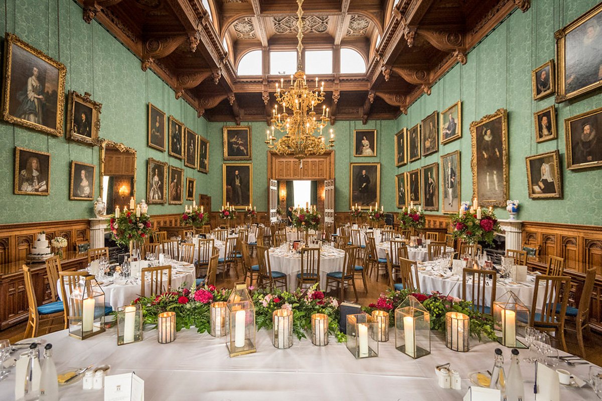 Elegant banquet hall with wooden paneling, framed portraits on green walls, ornate chandelier, and tables set for a formal event with floral centerpieces and candles.