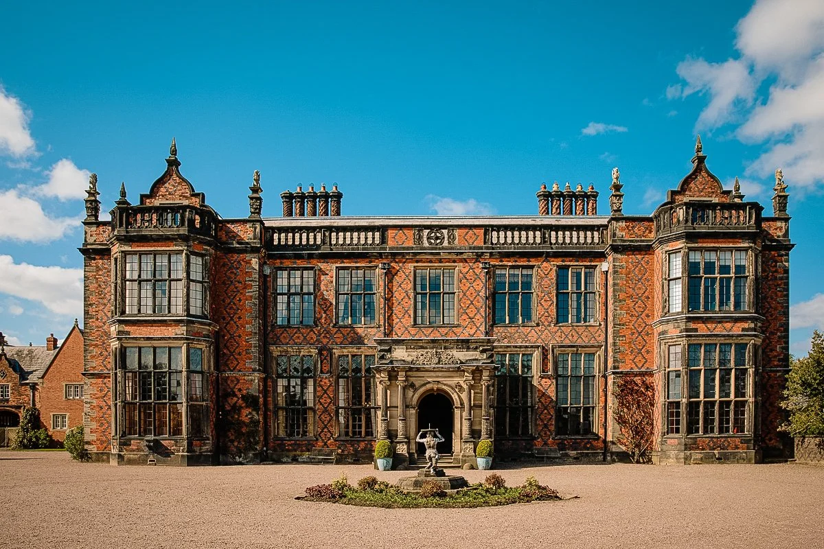 A large, historic brick mansion with ornate architectural details, multiple windows, and a central entrance with a stone archway, surrounded by a gravel courtyard and a fountain in front.