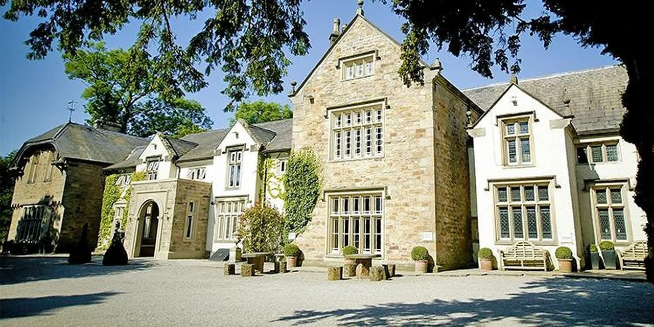 A large historic stone building with multiple windows, surrounded by trees with green foliage, and benches outside on a sunny day.