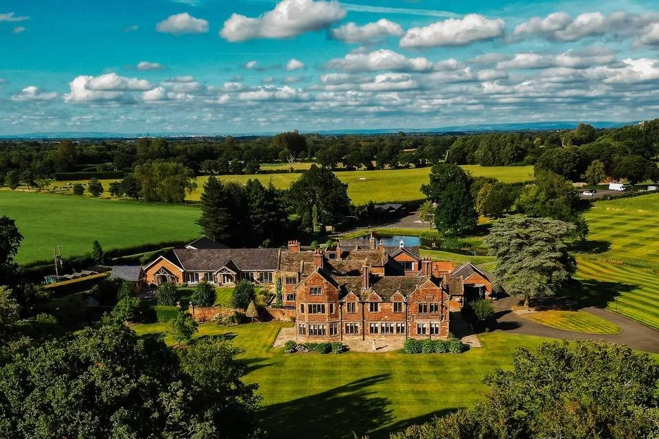 Large historic brick mansion surrounded by lush green lawns and trees, with expansive fields and farmland in the background under a partly cloudy sky.