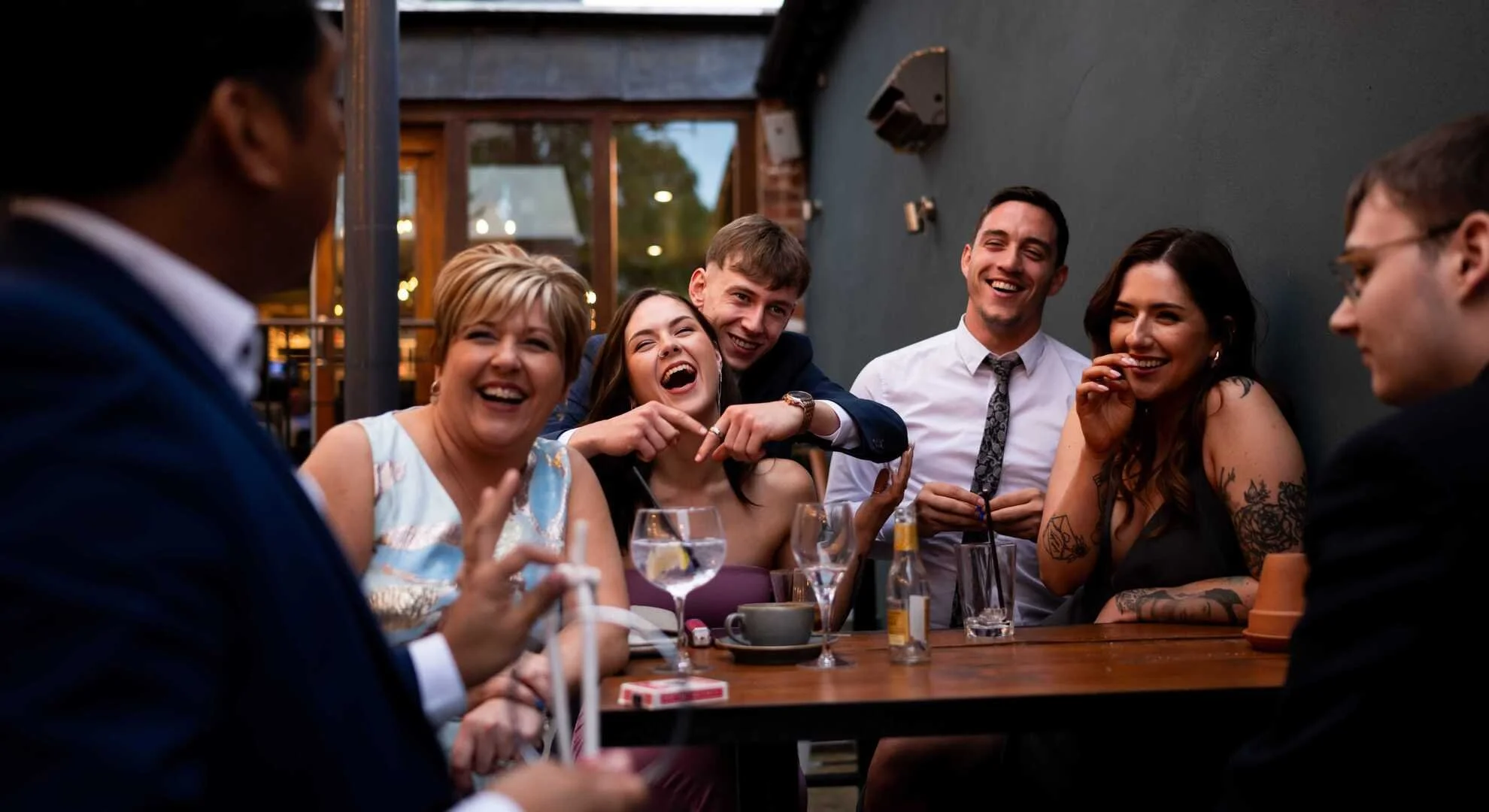 Group of people laughing and talking at an outdoor dinner party, sitting around a wooden table with drinks and snacks.