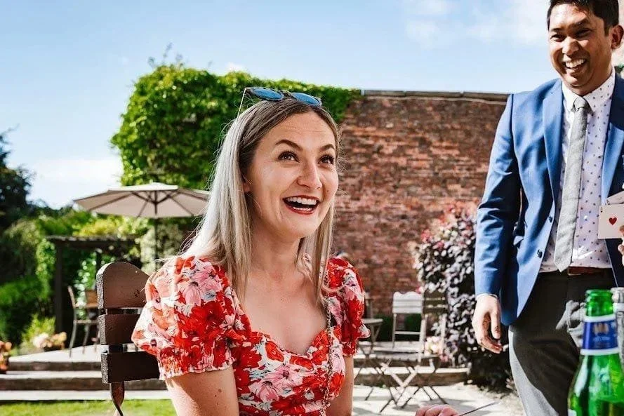 A woman with long blonde hair wearing a red floral dress, smiling excitedly, outdoors in a garden setting with a man in a blue suit next to her, holding playing cards.