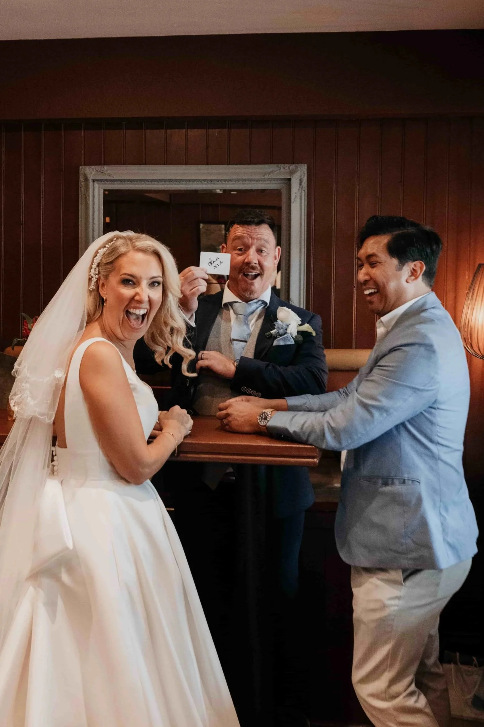 A bride in a white wedding dress and veil, smiling joyfully. A groom and another man, smiling and holding hands with the groom, are standing next to her in a warmly lit room with wood-paneled walls. The man in the middle is holding a small card with writing on it.