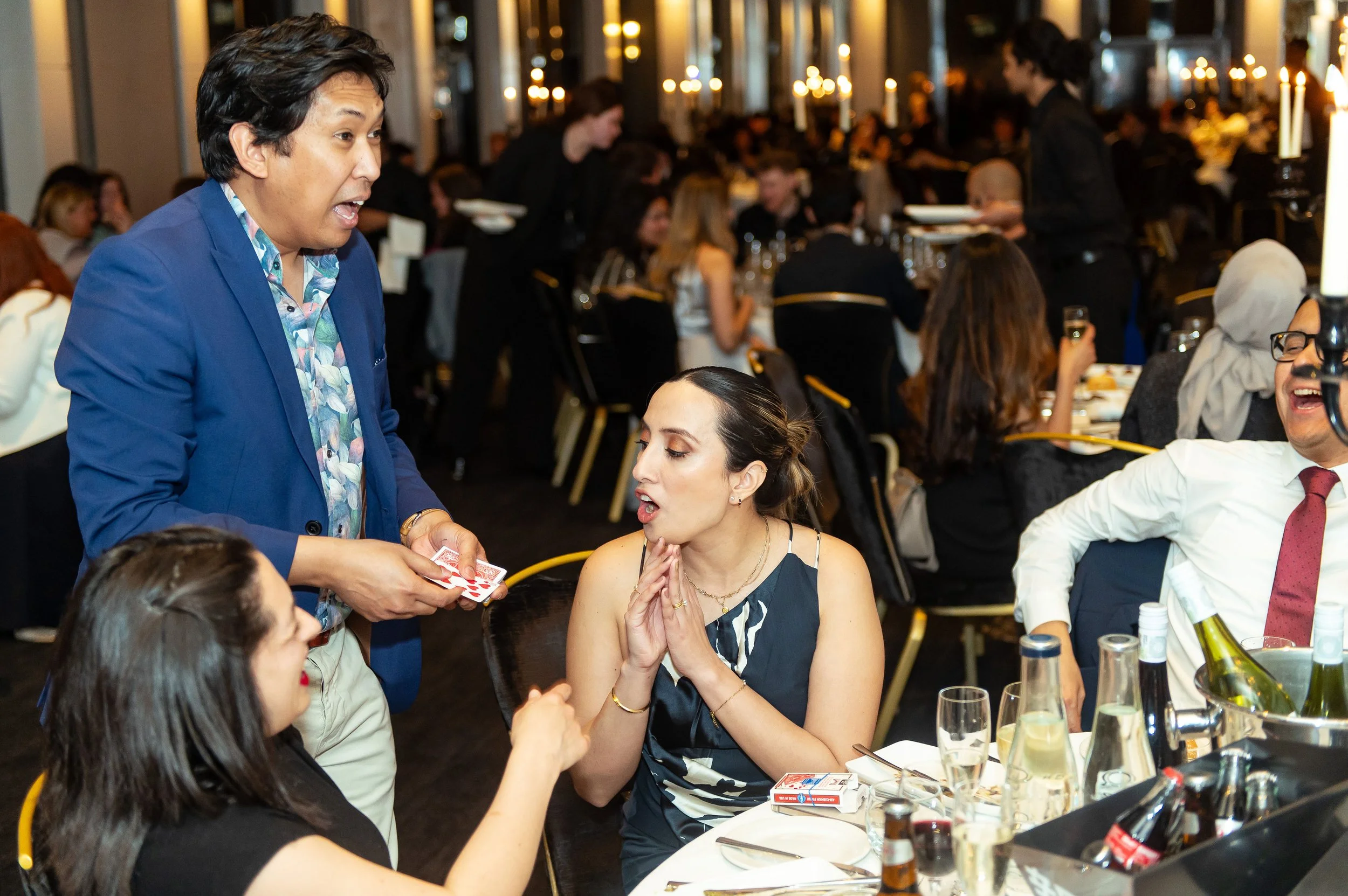 A man in a blue blazer and floral shirt performing a card trick for two women at a banquet or dinner event. The woman in the center is surprised and holding her hands near her face, while the other woman, facing away, watches intently. The scene is lively with many people seated at tables, and the room is decorated with candles.