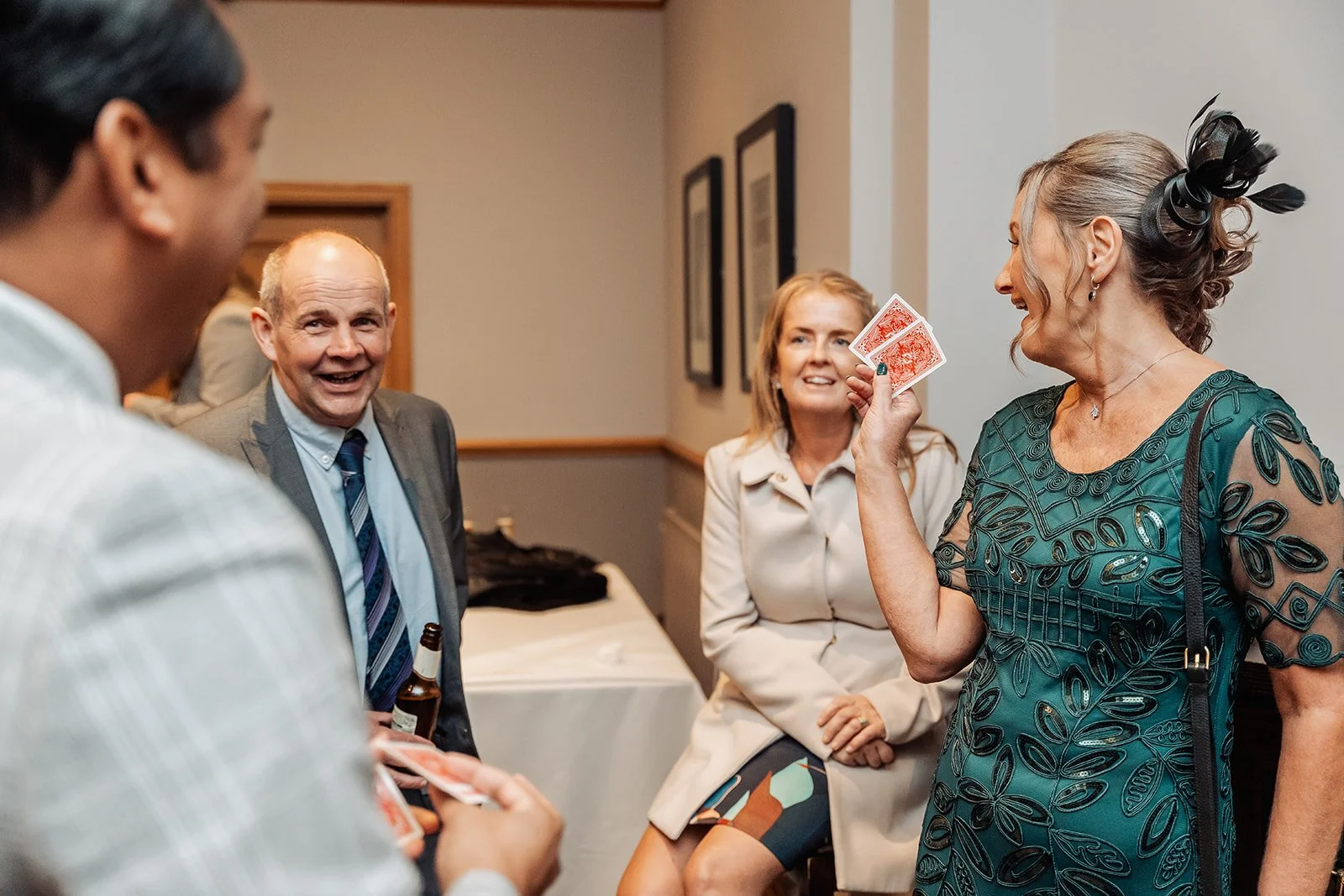 A group of four adults socializing indoors, with one woman holding playing cards and others holding drinks, engaging in conversation and smiling.