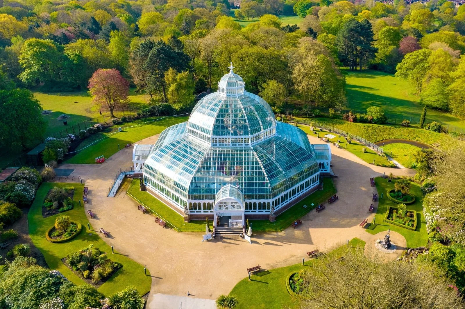 A large glass botanical conservatory or greenhouse surrounded by lush greenery and park-like grounds with walking paths, benches, and flower gardens.