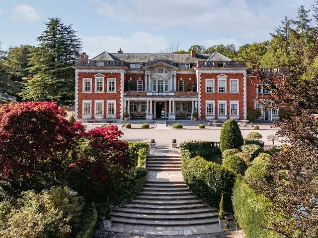 A large historic mansion with a brick facade, multiple windows, and a grand entrance, surrounded by well-maintained gardens and trees.