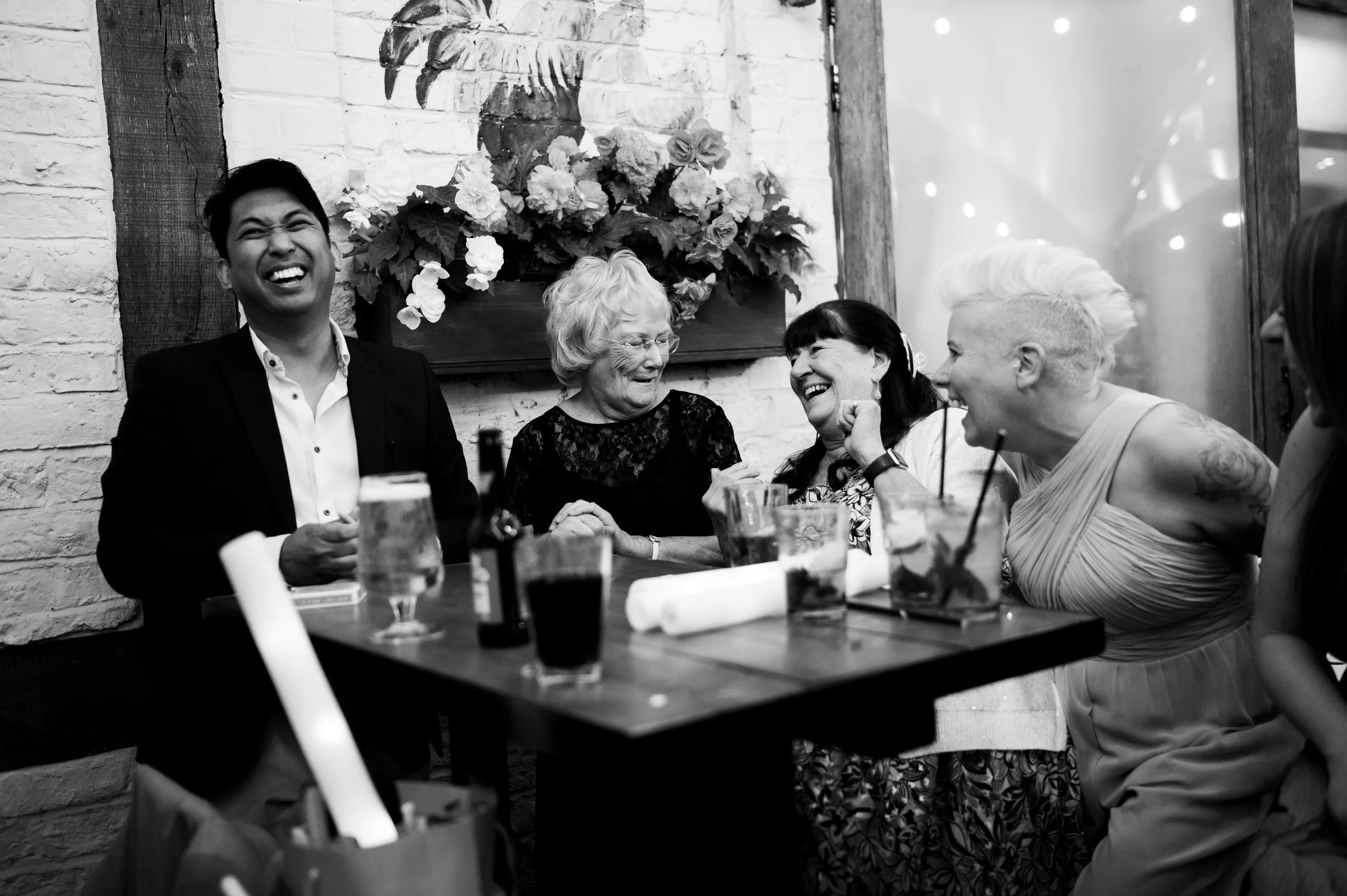 A group of five women and one man enjoying a lively conversation at a restaurant or bar, laughing and smiling, with drinks and bottles on the table in front of them. The setting has a rustic decor with a brick wall and floral art in the background.